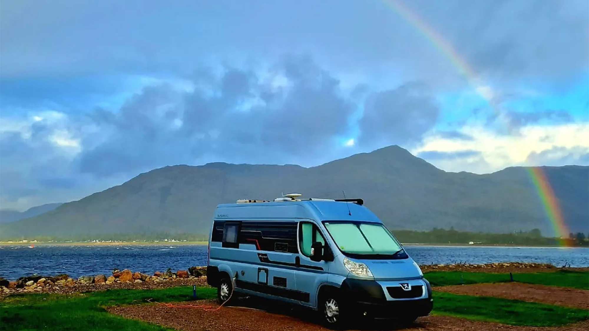 A motorhome parked on a pitch at Bunree campsite with the water behind leading to the far side where the mountains rise up under a blue but cloudy sky with a beautiful rainbow arching down on the water