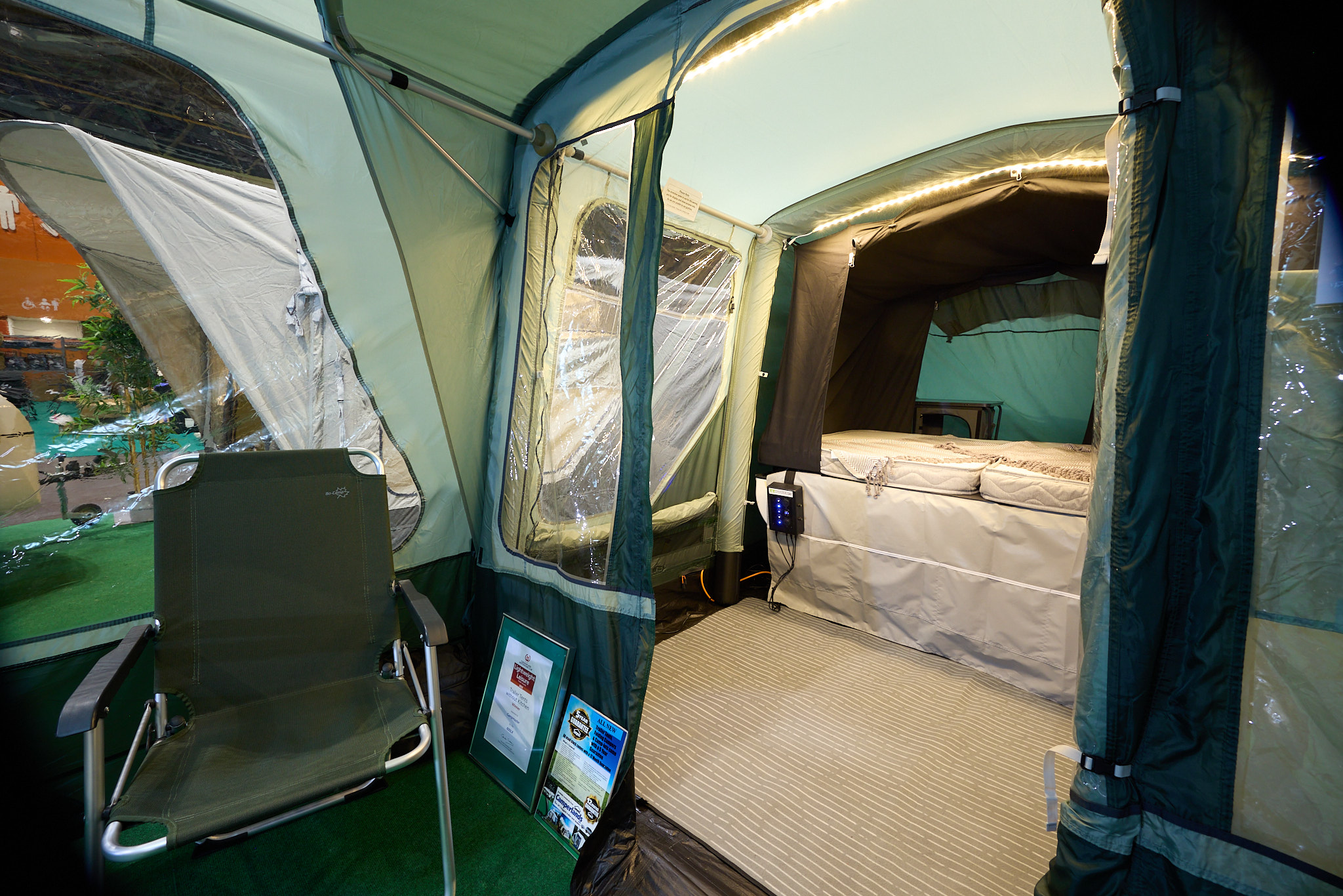Campmaster 600 Limited edition’s bedroom has a zipped door which is open revealing the bed.  There is LED strip lighting on the roof.  There is an indoor living area just outside the room with a beige groundsheet.  There's a green camping chair.