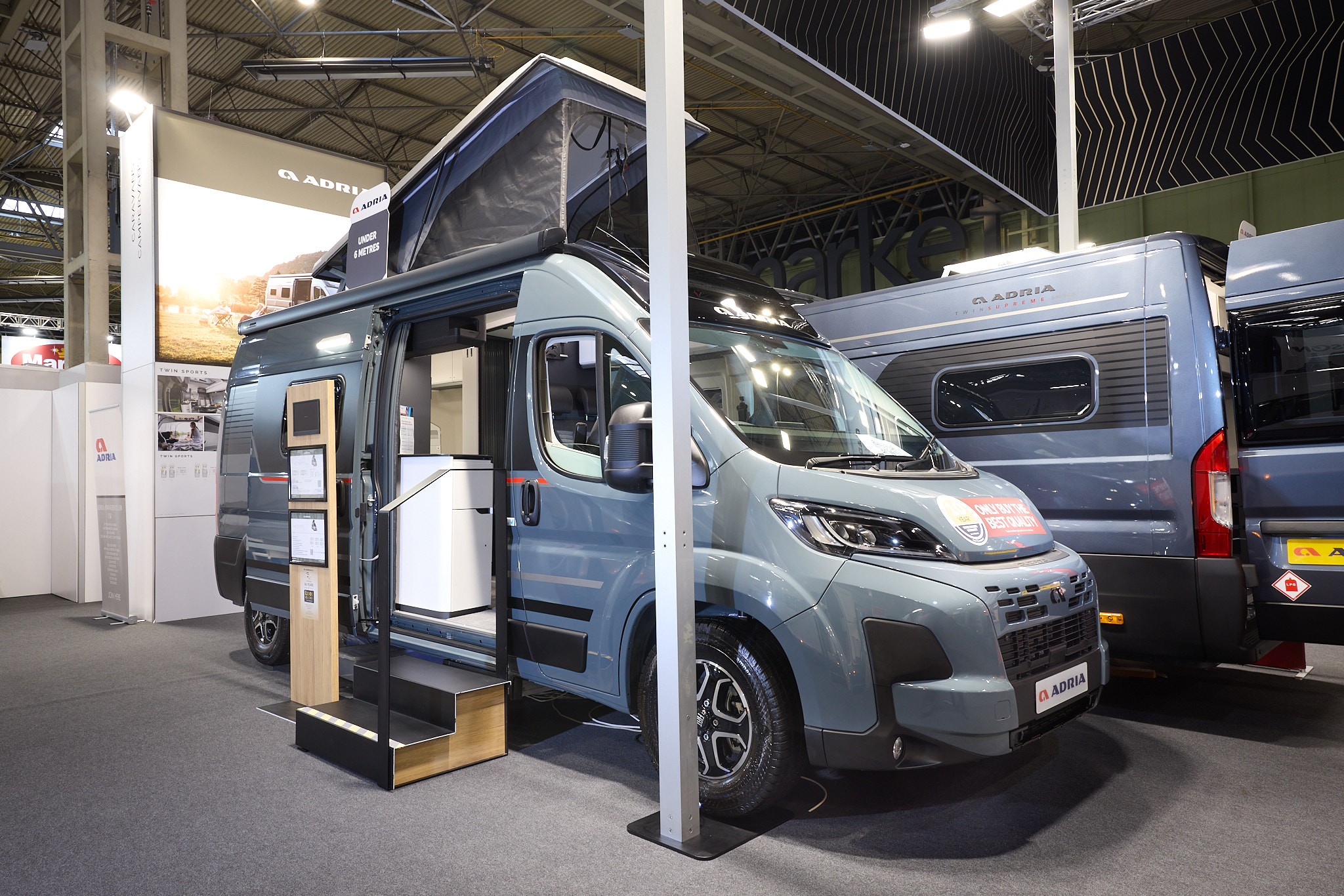 The Adria Twin Sport has a grey exterior.  The habitation door is open and there are two steps to gain easy access. There is an information board to the left of the steps.  The rising roof is open.
