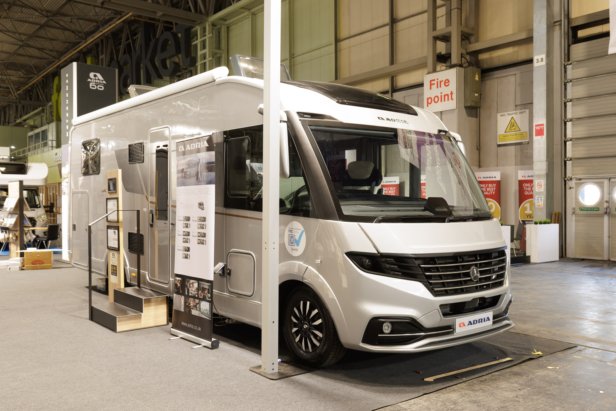 The Adria Supersonic 780 DL is silver.  There are two steps beside its habitation door.  There is a roller banner one side of the steps with an interaction stand on the other side.  Its skylights are open.