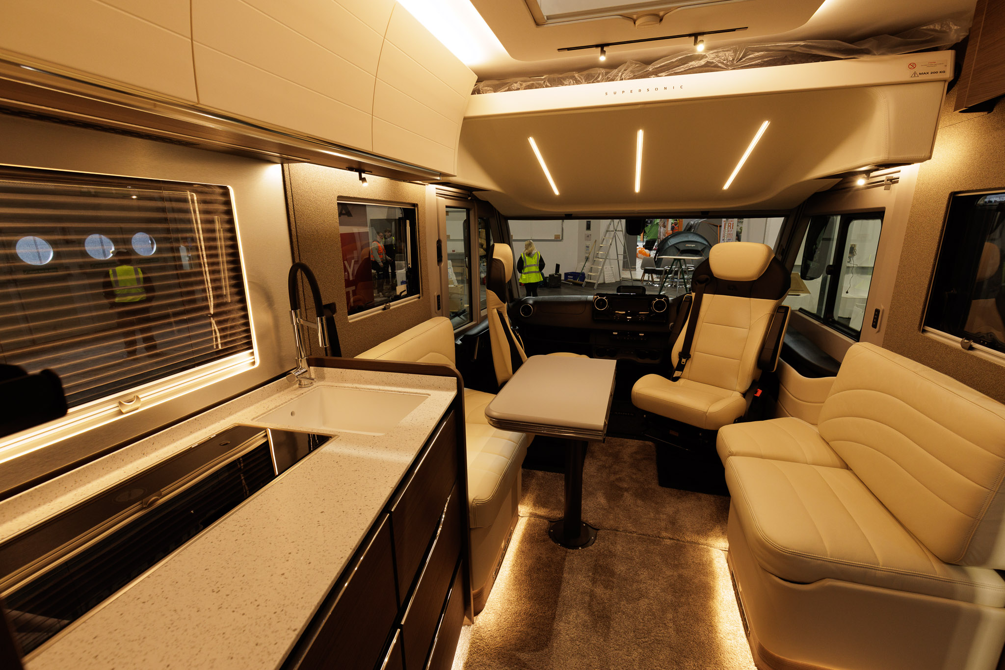The Adria Supersonic 780 DL has cream upholstery and the front seats are swivelled.  There is a table which is folded but can be extended.  There is a roof bed above the cab.  The kitchen is to the left.