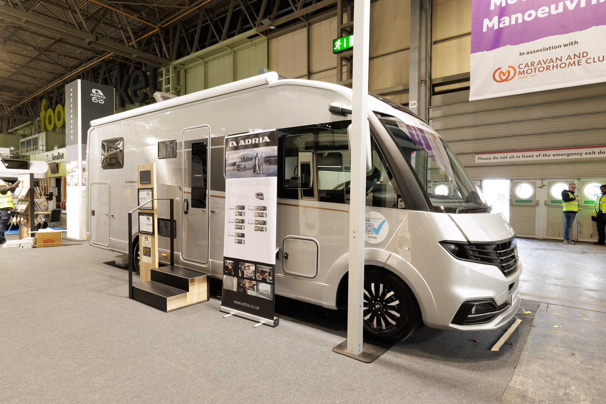 The Adria Supersonic 780 DL is silver.  There are two steps beside its habitation door.  There is a roller banner one side of the steps with an interaction stand on the other side.  Its skylights are open.