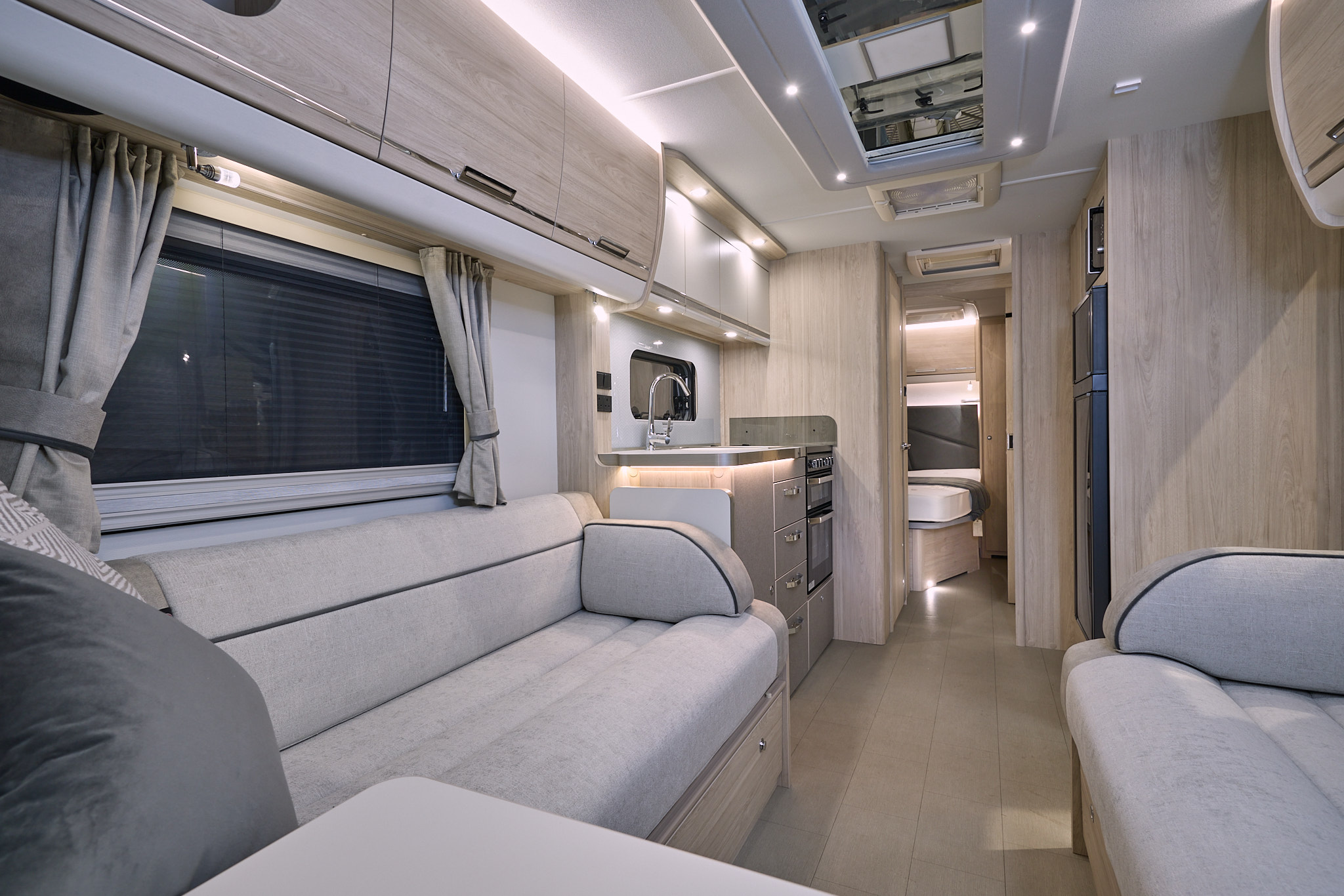 Elddis Crusader Tempest’s interior has grey upholstery with dark grey accents.  It has pale wooden furniture.  The kitchen is in the middle and the bedroom door is open revealing a fixed French transverse bed.