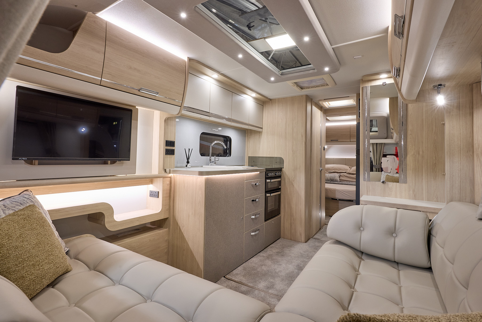 Interior of the Elddis Crusader Borealis with cream leather sofas, cosy cushions, wood accents and beige carpets.  The door to the rear bedroom is open.  Perfect for relaxation and designed for comfort.