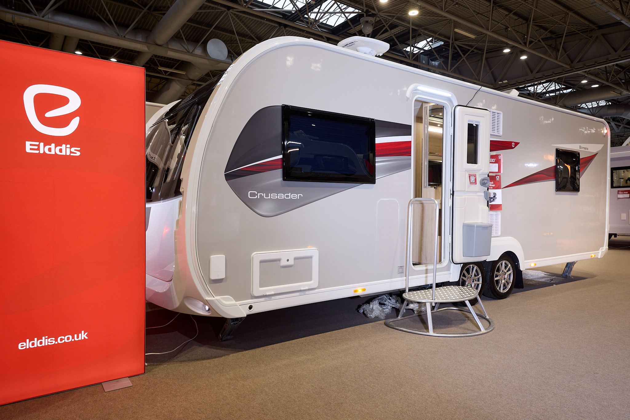 The Elddis Crusader Borealis has a grey exterior with grey, red and black decals.  The entrance door is open and there is a metal step to gain easy access.  There is a large Elddis red banner with their website address next to it.