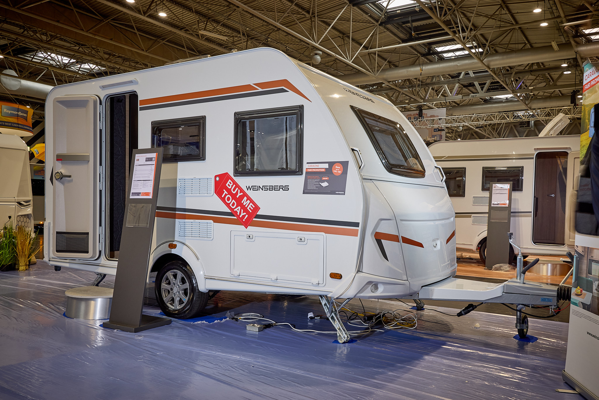 Weinsberg CaraOne 390 QD is white with orange and black decals.  Its entrance door is open and there is a metal step to gain easy access.  There is an information board next to the step.