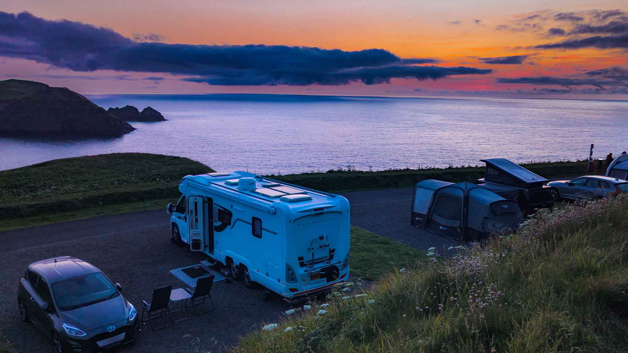 Looking down from a grassy hill out over the motorhome on the pitch next to the sea under an orange sky fading into the night