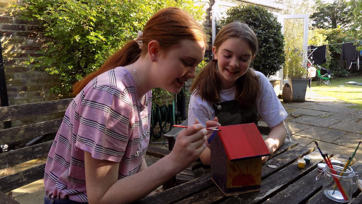 Two girls painting a birdbox on a patio in a back garden.