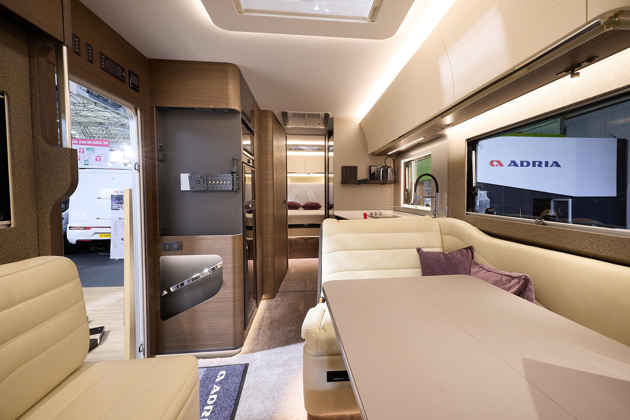 Adria Supersonic's interior has cream upholstery with curved dark wooden units.  There are two dark pink cushions, it has a sky light and a beige table.  The fixed bed is to the rear and there is a place for a TV on the left hand side.