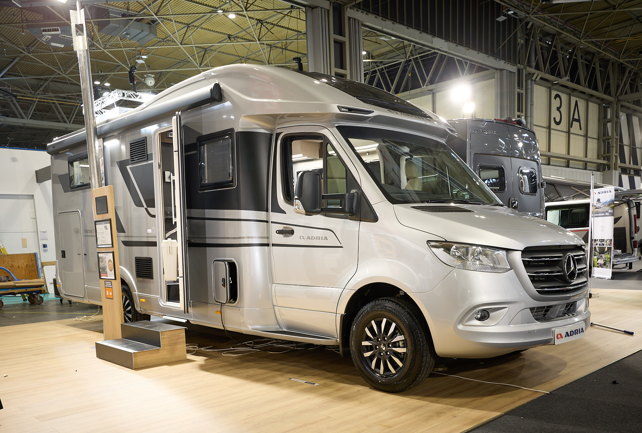 Adria Matrix Supreme's exterior is silver and black, the entrance door is open and there are wooden steps to gain easy access.  There is an interactive information board by the side of the van.