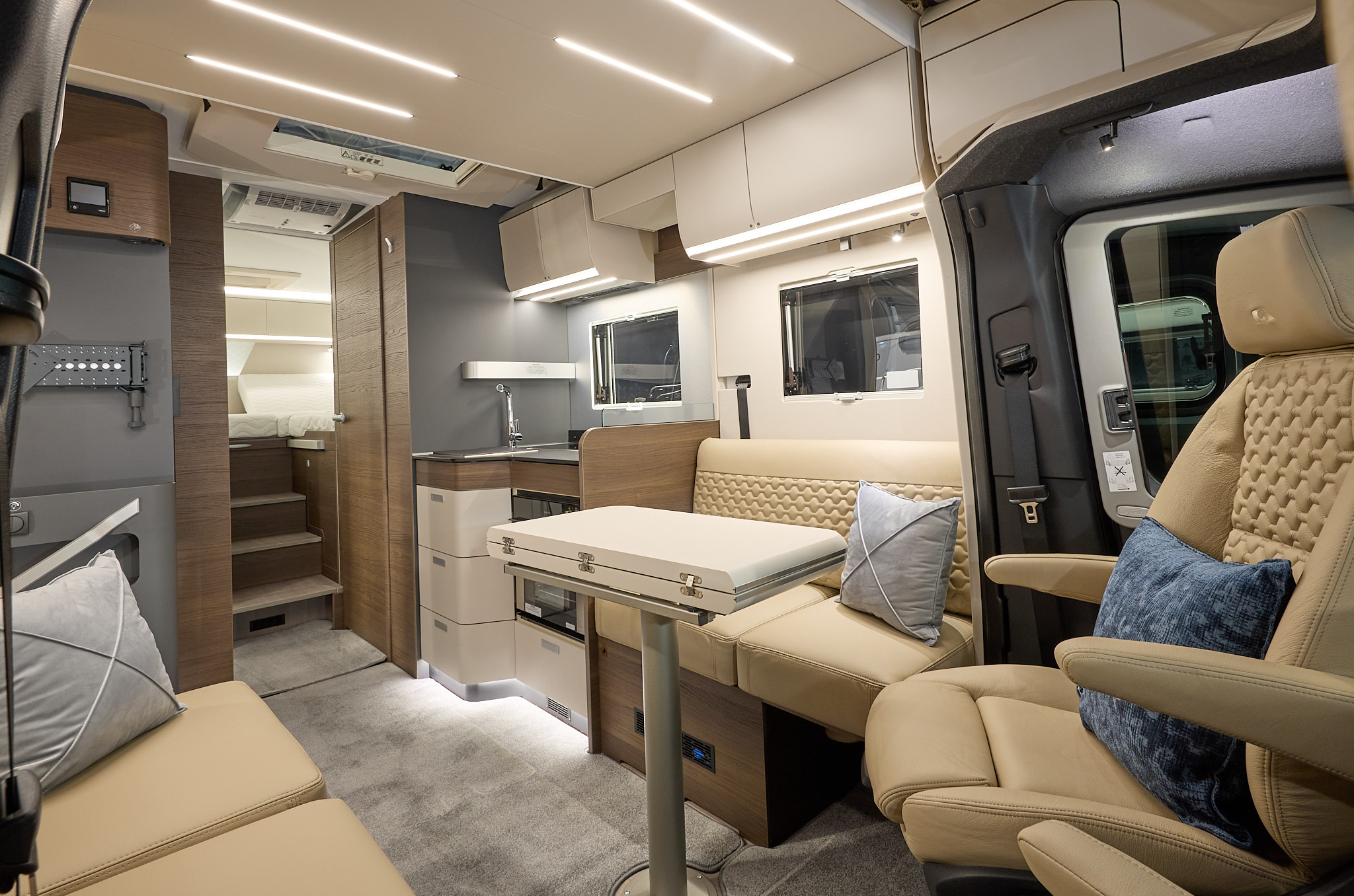 Adria Matrix Supreme's interior has beige leather upholstery with wooden units.  There are two blue cushions, it has a cream ceiling and a cream table which is folded.  The steps to the fixed bed are to the rear and the TV station is on the left.