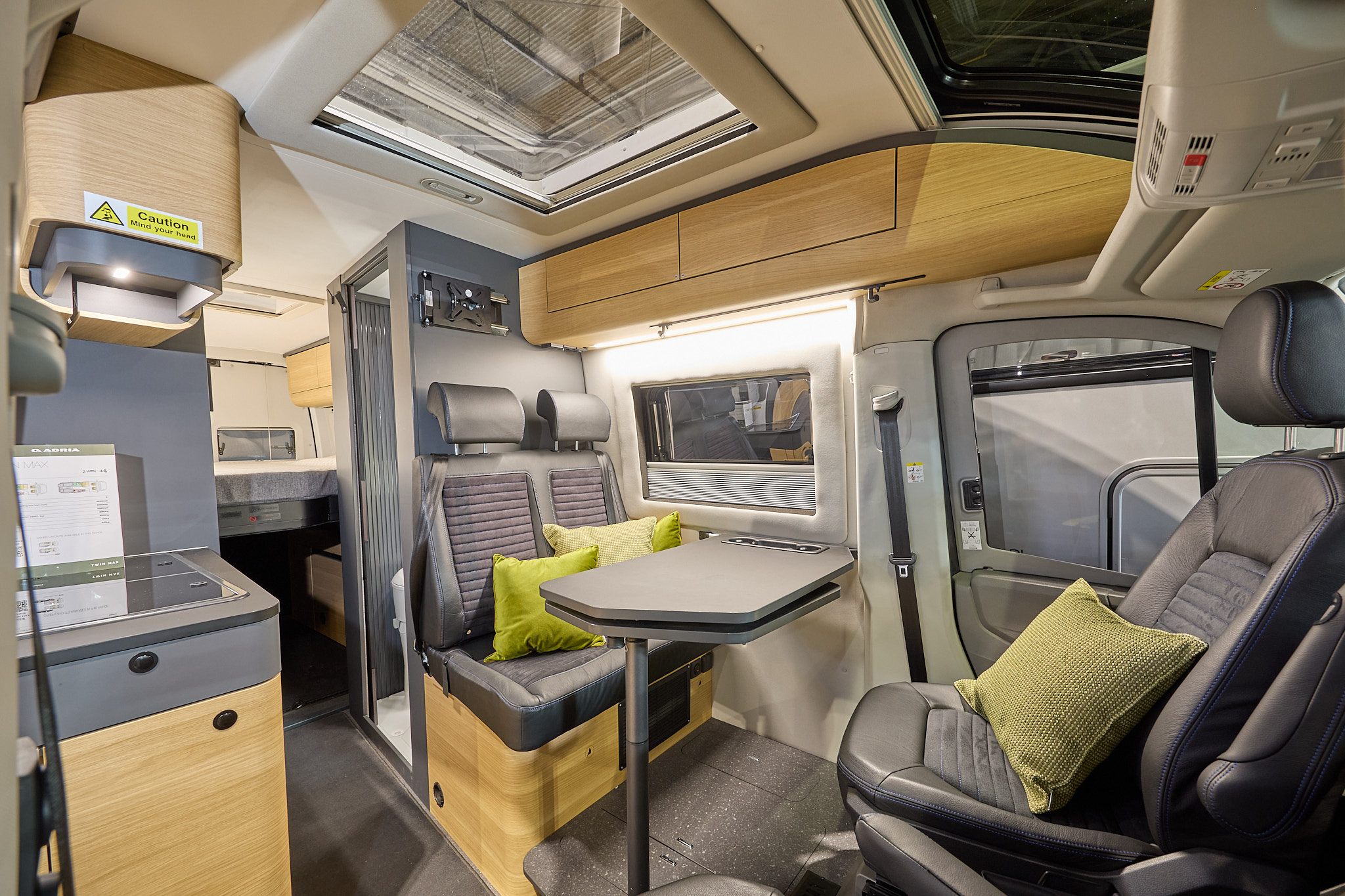 Adria Twin Max's interior has brown upholstery with curved wooden units.  There are four lime green cushions, it has a clear sunroof and the driver’s and front passenger's seat are swivelled.  The fixed bed is to the rear with the hob on the left.