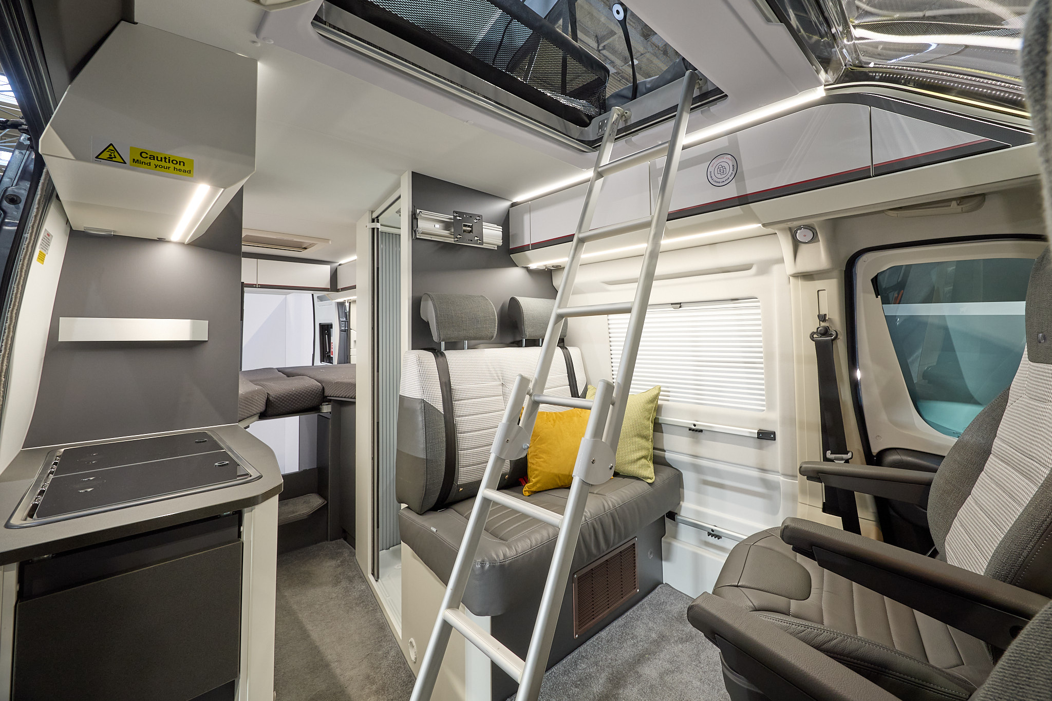 Adria Twin Sport's interior has brown and beige upholstery with two yellow cushions, there is a silver ladder to the roof bed and the driver’s and front passenger's seat are swivelled.  The fixed bed is to the rear with the hob on the left hand side
