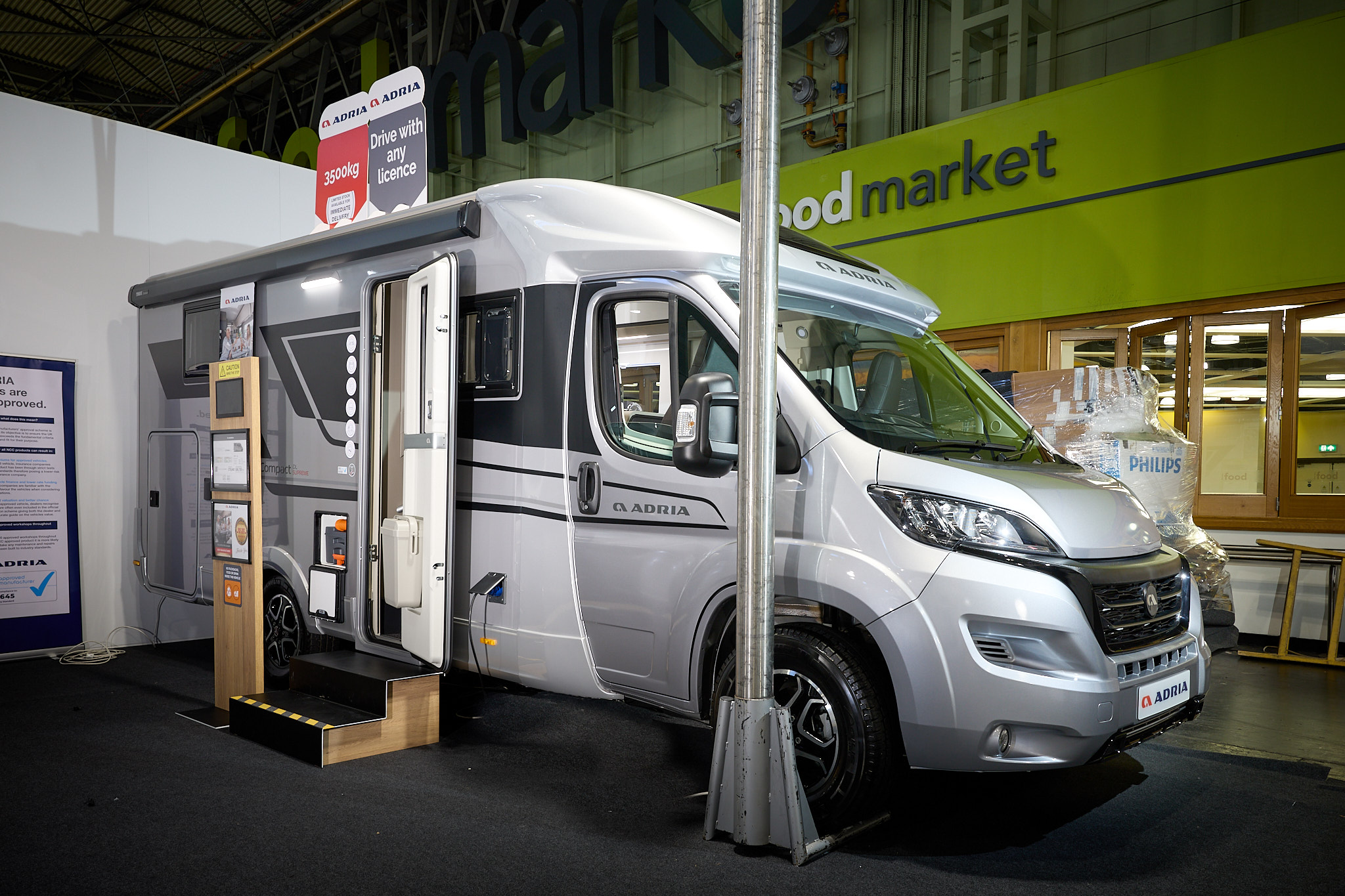 Adria Compact Supreme's exterior is silver, white and black, the entrance door is open and there are wooden steps to gain easy access.  There is an interactive information board by the side of the van.   