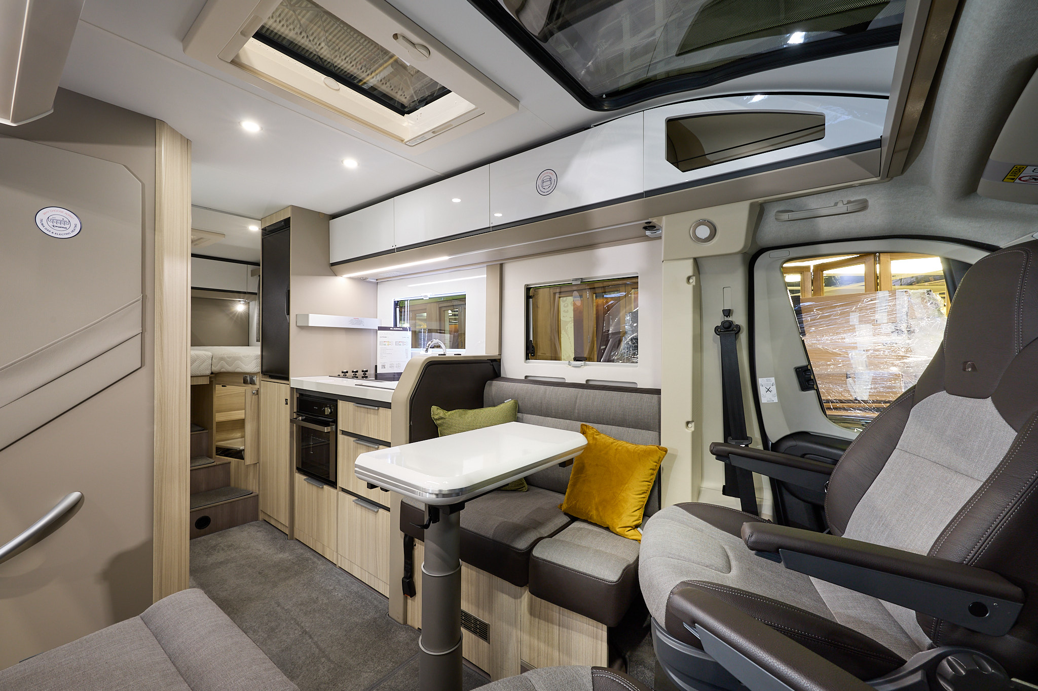 Adria Compact Supreme's interior has beige and grey upholstery with pale wooden units.  There are two yellow cushions, it has a large sunroof and a folded cream table.  The steps to the fixed bed are to the rear and the two front seats are swivelled.