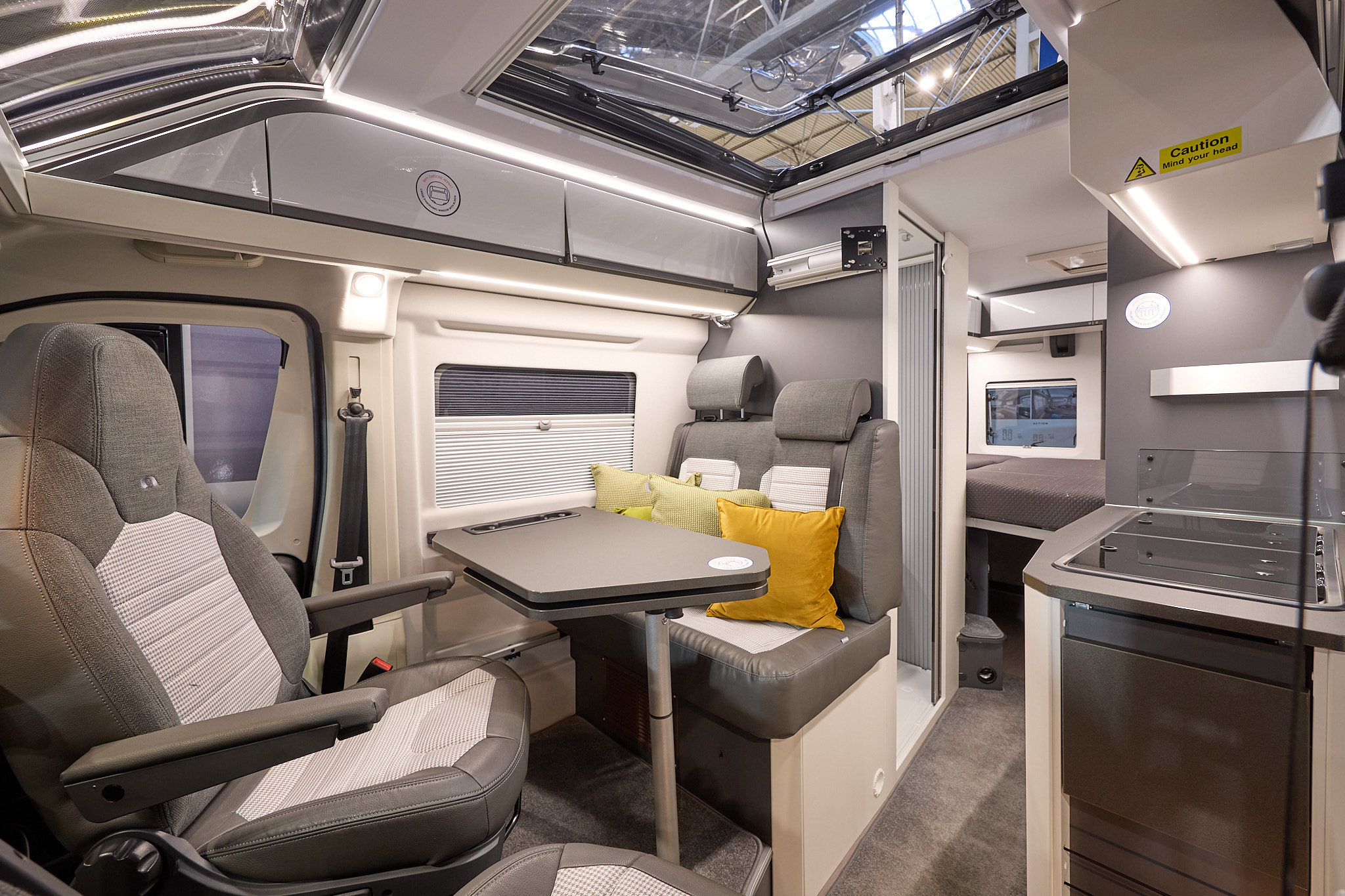 Adria Twin Supreme's interior has brown and beige upholstery with three yellow cushions, there is a large clear sunroof and the front passenger's seat is swivelled.  The fixed bed is to the rear with the hob on the right hand side.