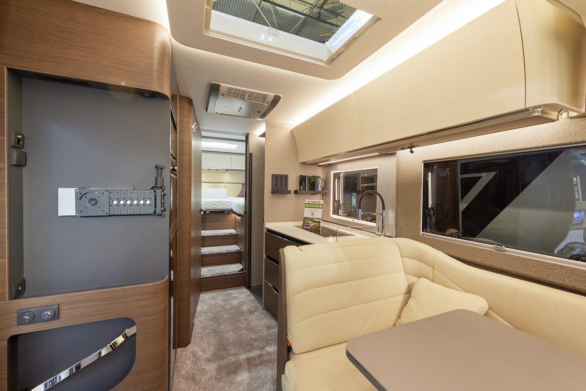 Adria Supersonic 780 SL interior which has cream upholstery, with cream overhead cabinets and wooden curved panelling.  The carpet is mid brown, there are three steps leading to the fixed rear bed.  There is a large roof light above the kitchen.