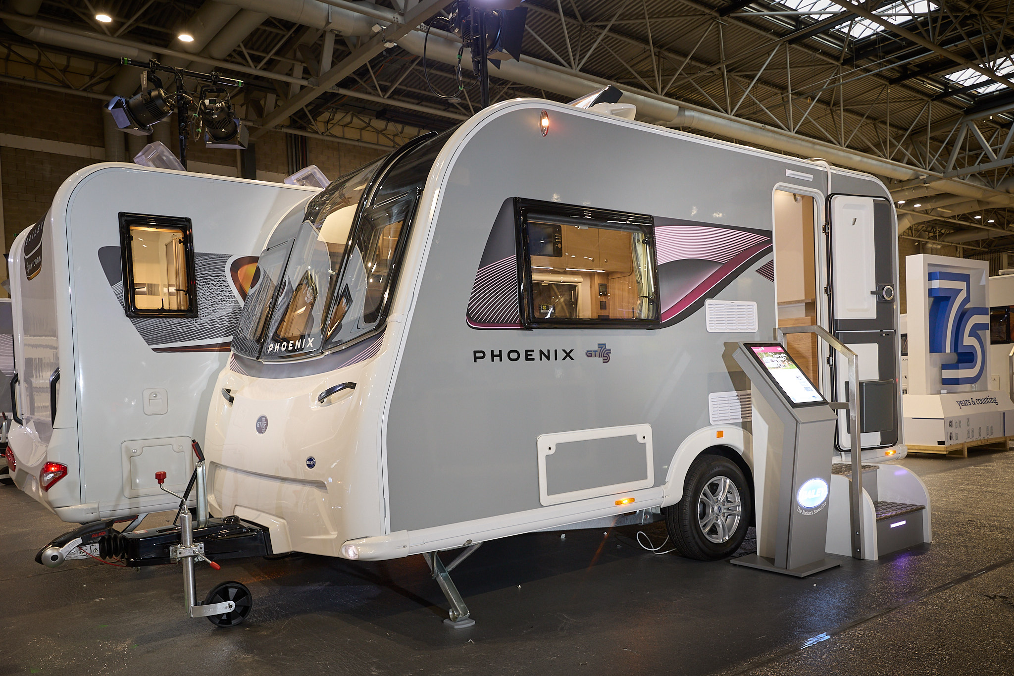 Bailey Phoenix GT75 420 exterior which has pale grey sides with an all white front.  The door is open and there are steps to gain easy access to the interior.  The skylight is open and there is an interactive pedestal next to the steps