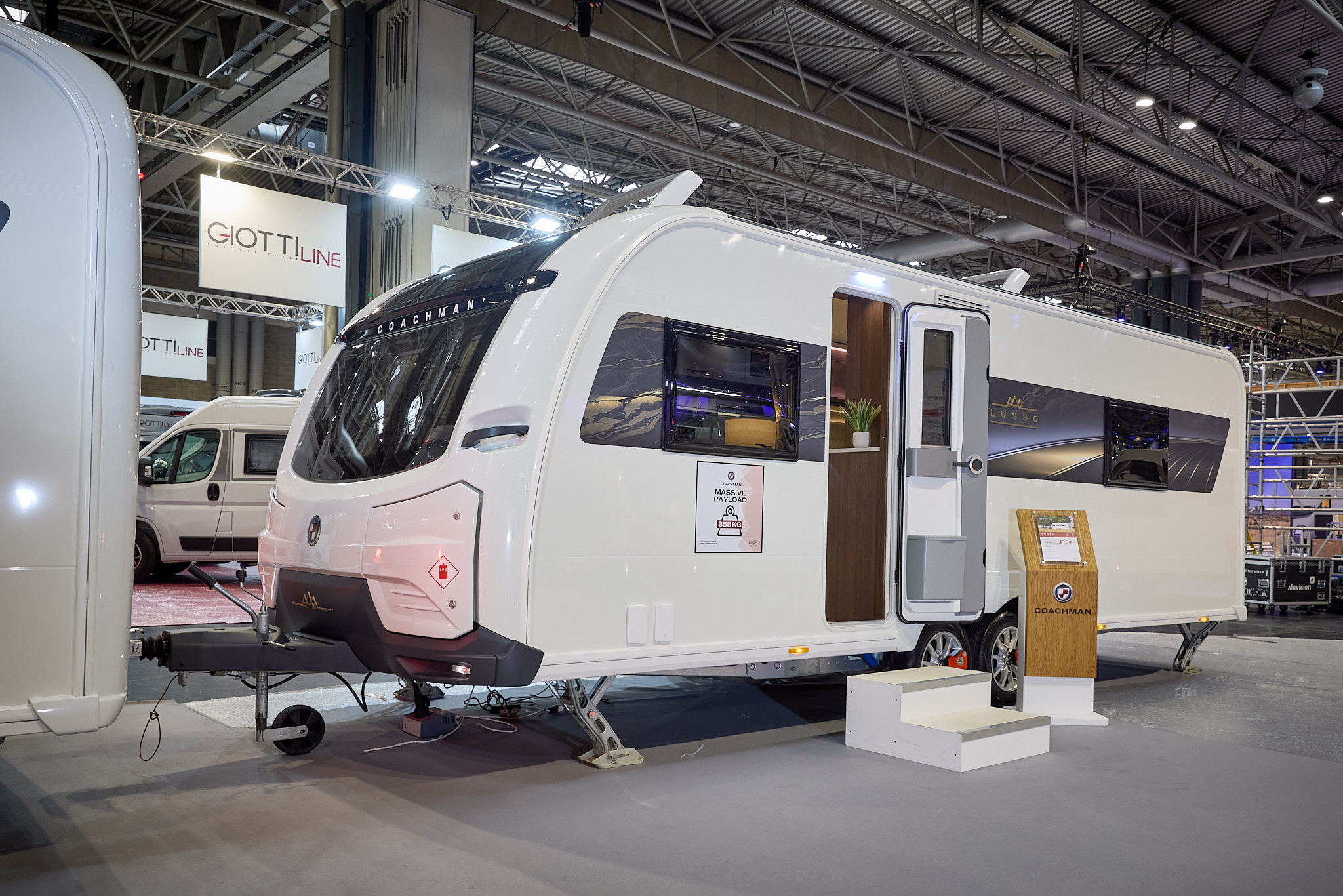 The Coachman LUSSO II has a white exterior with a brown patterned decal.  Its sunroof is open.  The entrance door is open and there are two wooden steps to gain easy access.  There is a stand next to the steps.