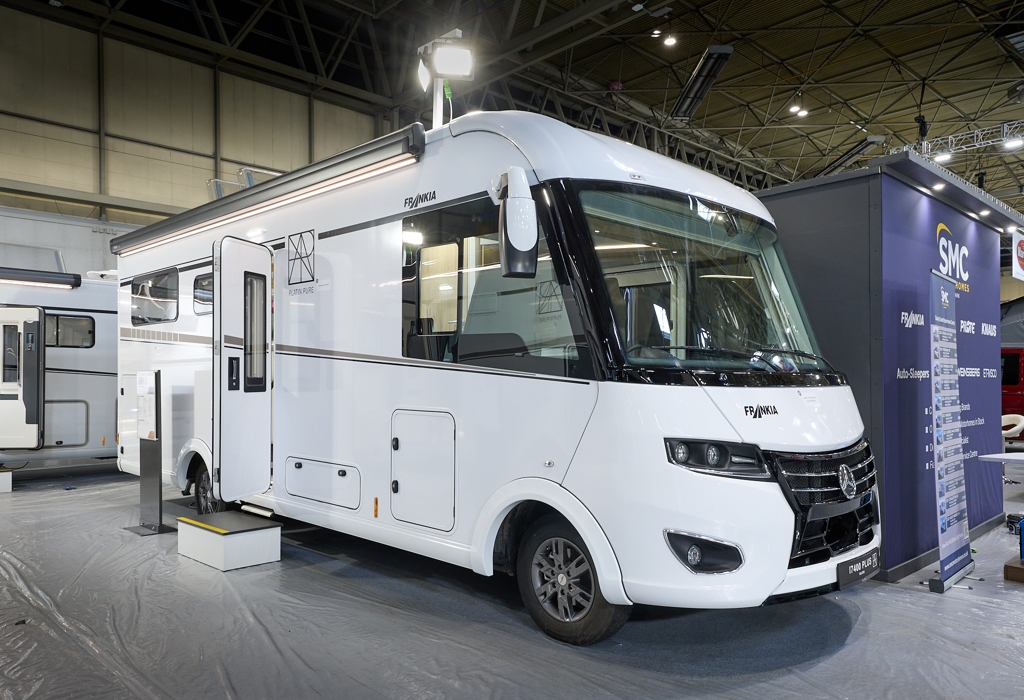 Frankia I 7400 PLUS Platin Pure Edition’s exterior is white.  Its entrance door is open.  There is a step to gain easy access with an information stand next to it.  