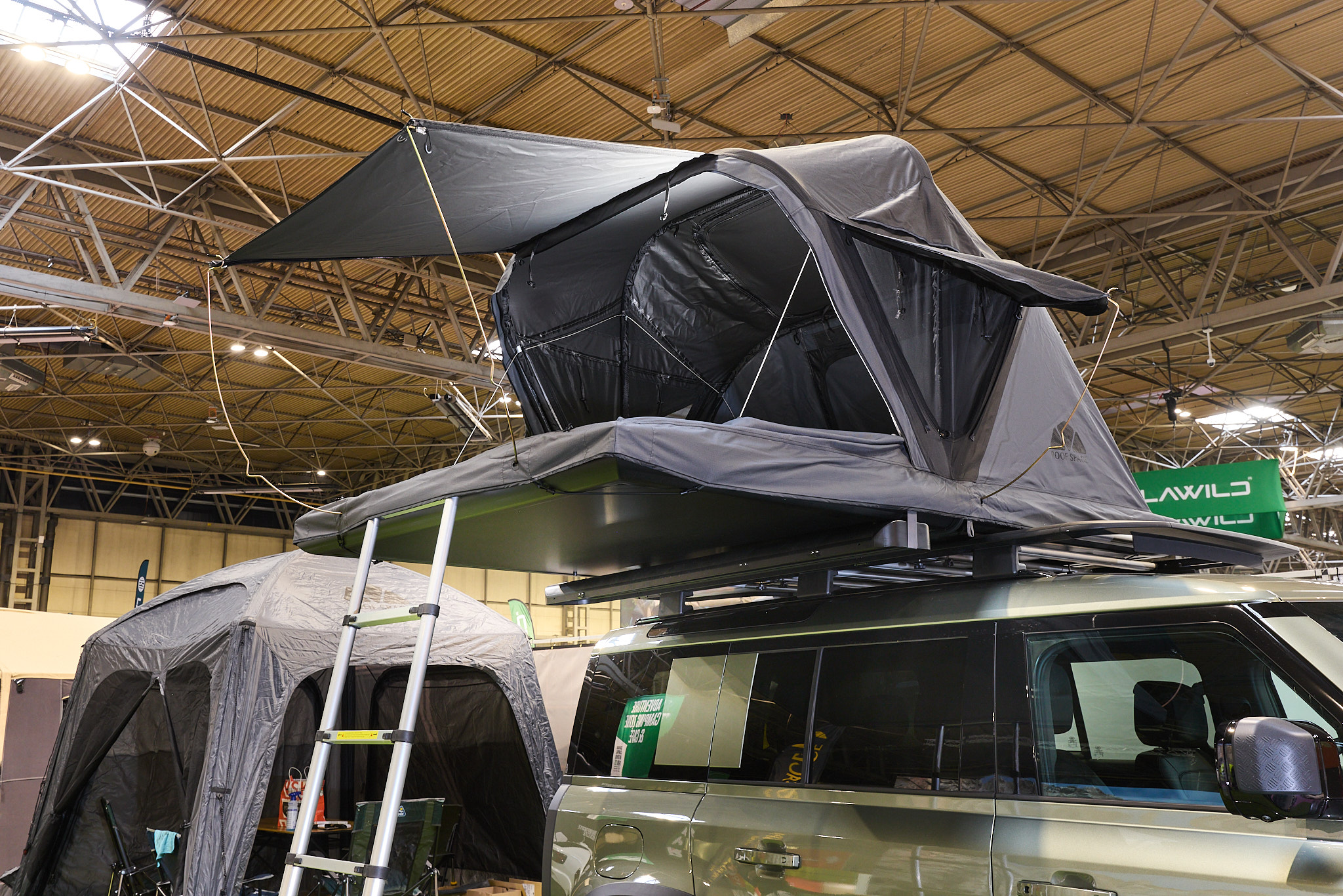 The Roof Space 4 is fully extended on a Land Rover Defender.  The canvas is pale grey, with a black porch.  There is a platform with a sturdy ladder to gain easy access.