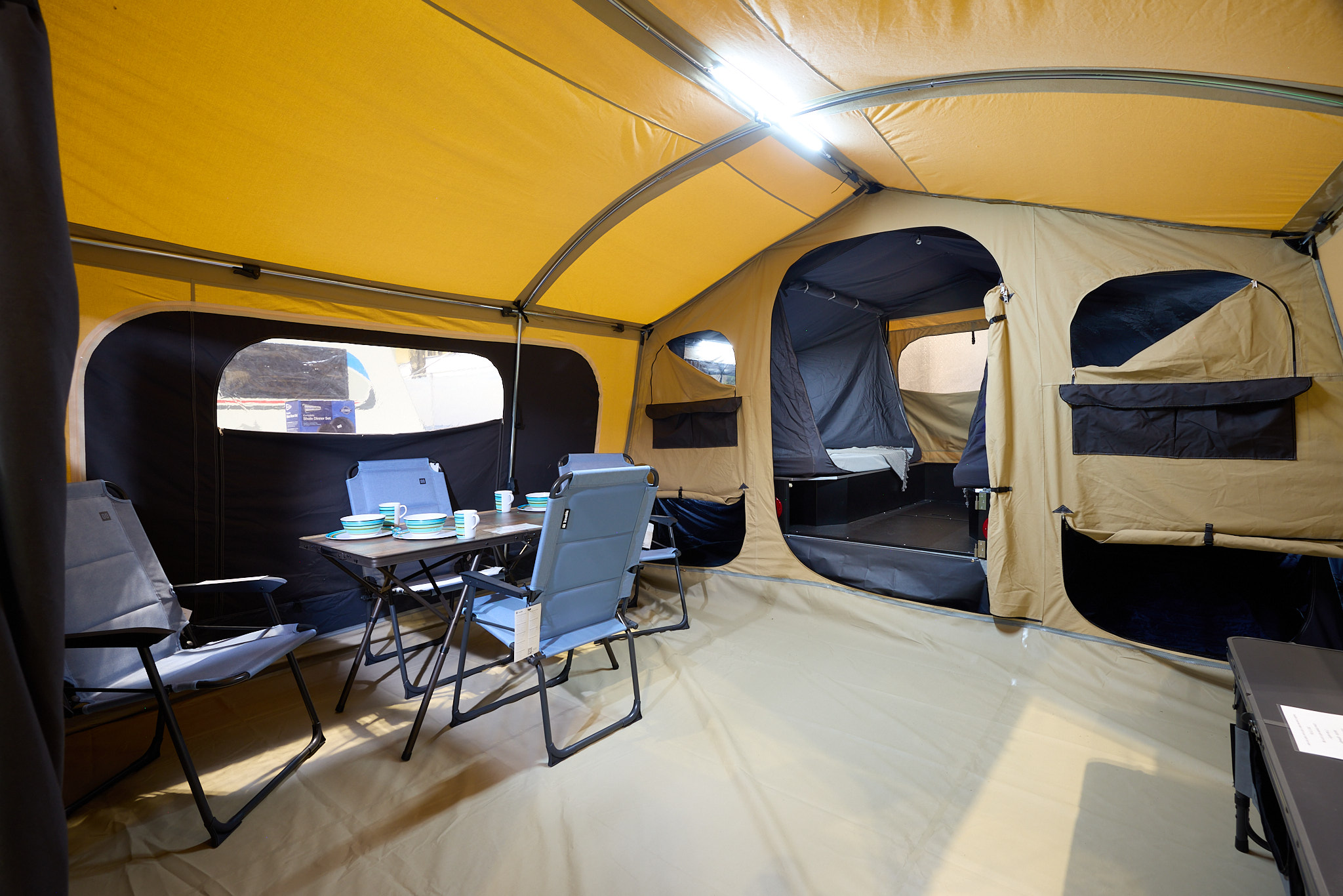 The Trigano Camplair S has yellow coloured canvas with a cream room divider.  The bedroom door is open.  There is a step to the bedrooms.  There is a light on the ceiling.  There is a dining table with four blue chairs