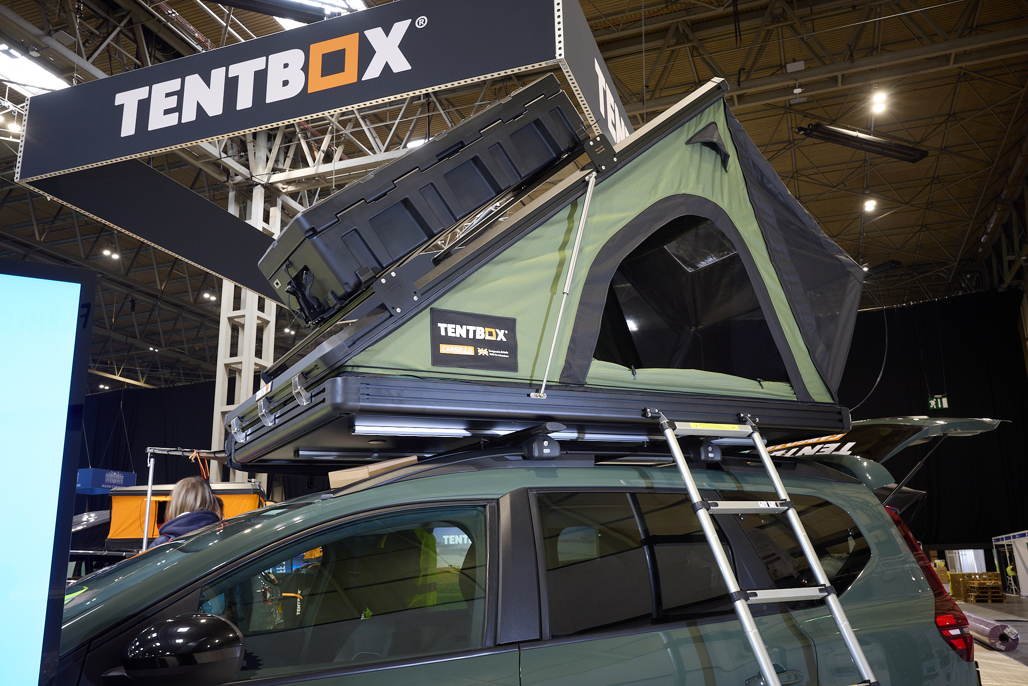 TentBox Cargo 2.0 is shown on a Dacia.  The canvas is green and it has a black hard top with a storage box.  There is a silver ladder to gain access.
