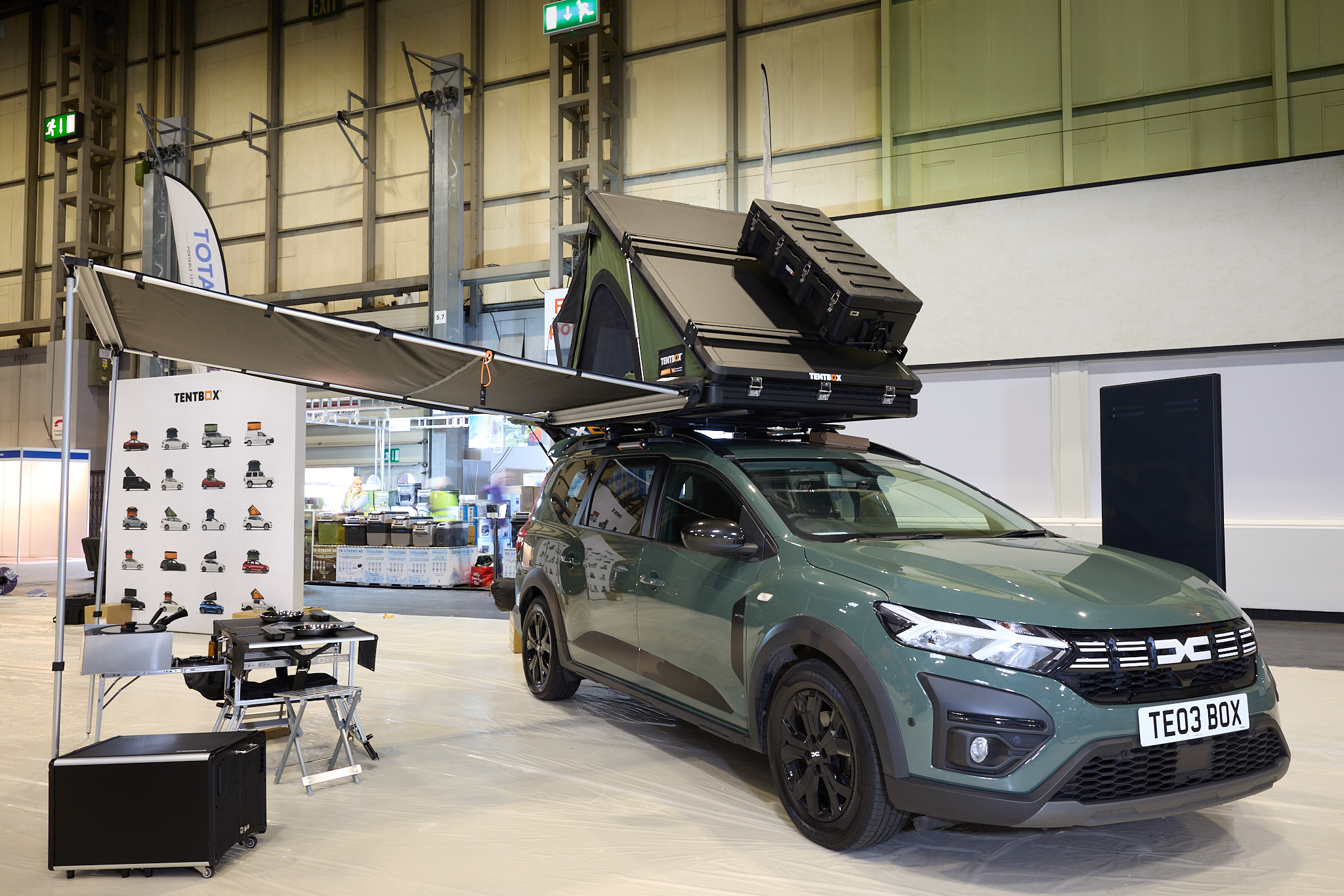 TentBox Cargo 2.0 is shown on a Dacia.  The canvas is green and it has a black hard top with a storage box.  There is an awaning and there are table and chairs underneath.