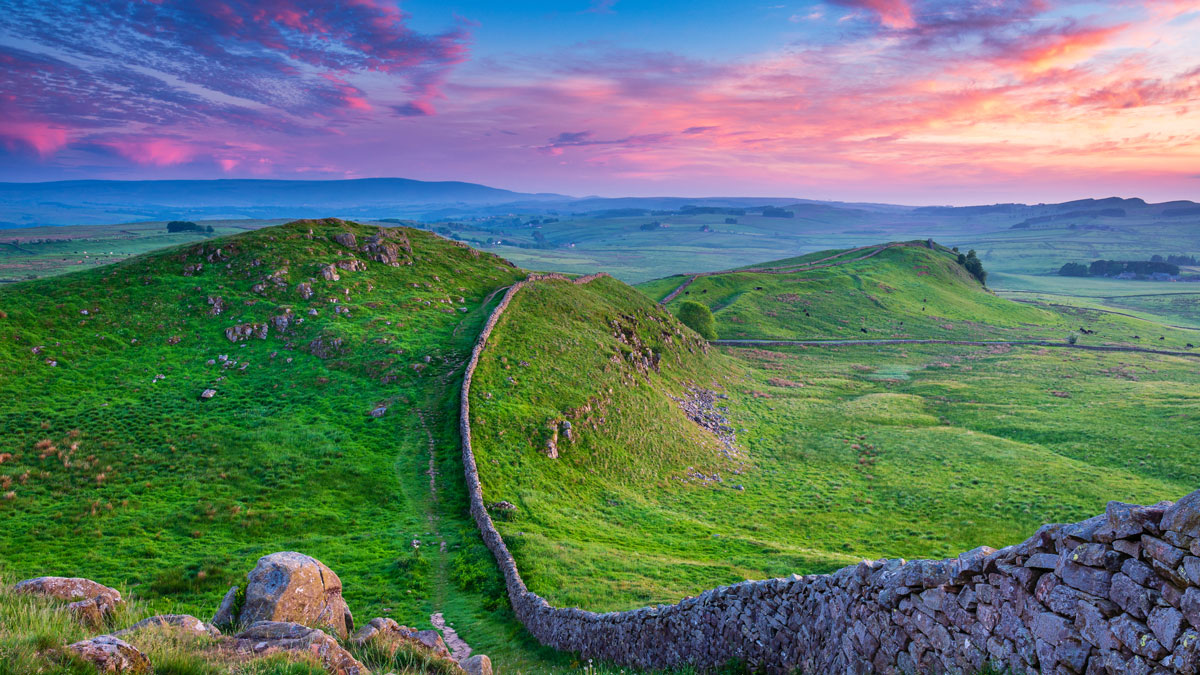 Hadrian's wall rising up and down over the green hills with a beautiful blue and pink sky in the backdrop