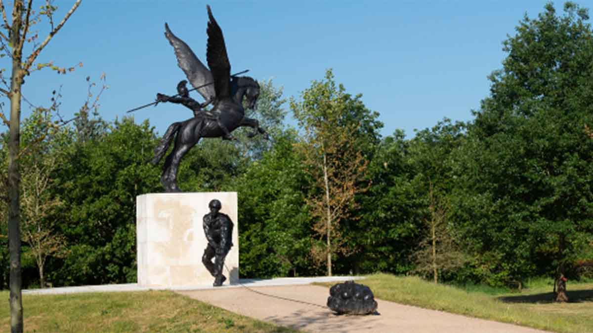 The National Memorial Arboretum. The statue of a solder in front of a white brick monument with the sculpture of a horse with wide wings. The memorial is surrounded by tall gree trees under a blue sky