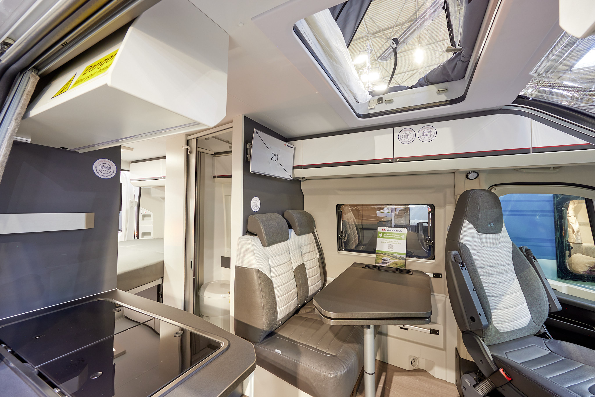 Adria Twin Sports 600 SPB interior, rear passenger seats in brown and cream, fixed table, swivelled driver’s seat which can double as a dining chair.  Also shown is a hob, access to the rising roof and the combined washroom and toilet.  