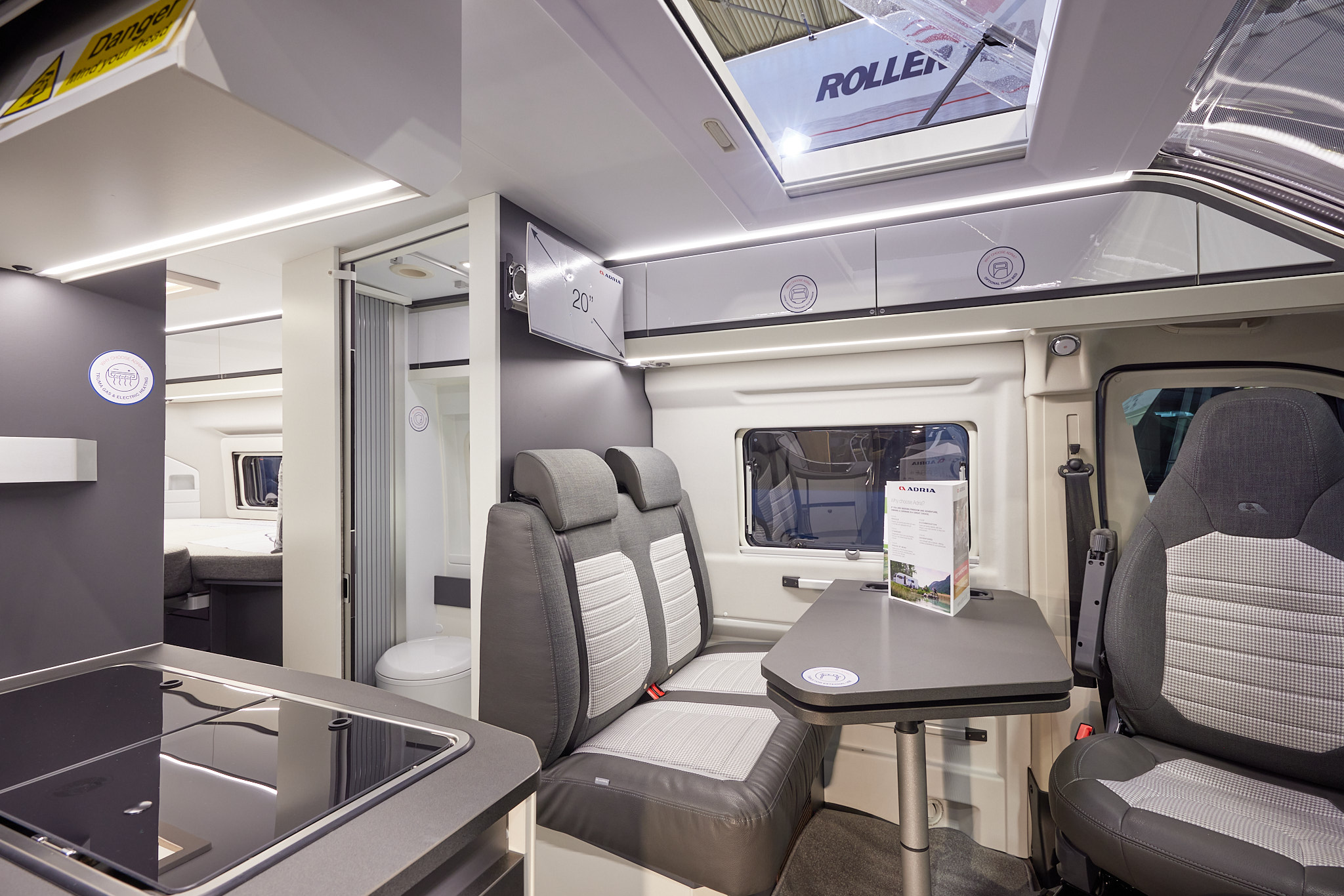 Adria Twin Supreme 640 SLB interior, rear passenger seats in brown and cream, fixed table, swivelled driver’s seat which can double as a dining chair.  Also shown is a hob, an open roof light and the combined washroom and toilet.  