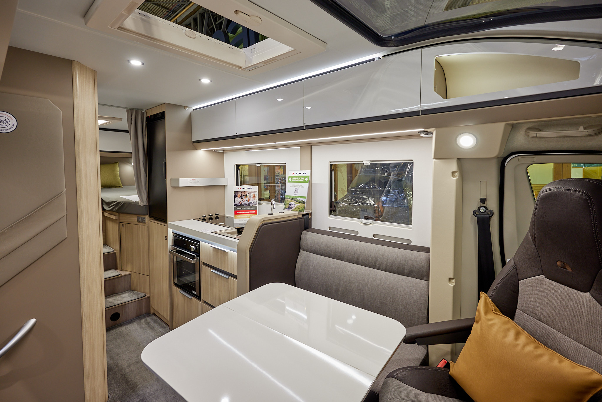Adria Compact Supreme DL has grey upholstery, with shiny white overhead cabinets and table.  It has light wooden panelling, there are mid-brown carpets, there are three steps leading to the fixed rear bed with a privacy curtain.