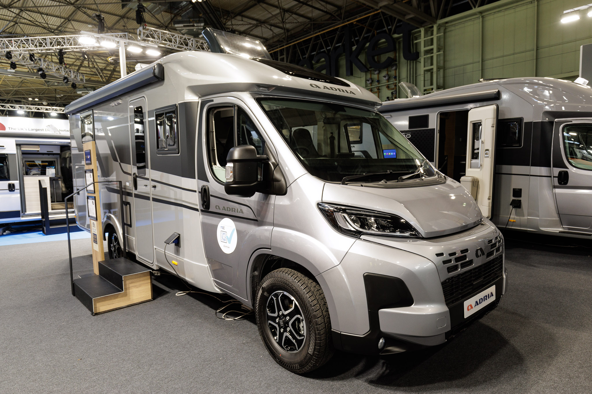 The Adria Compact Supreme DL is silver with a white coachbuilt body.  It has dark grey decals along its side.  There are two steps up to the main door to enable easy access.  It has an interactive stand by the side of the steps.