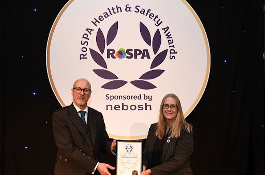 Global health and safety award winners. Two people  holding a certificate and smiling for the camera in front of The Royal Society for the Prevention of Accidents (RoSPA)  image