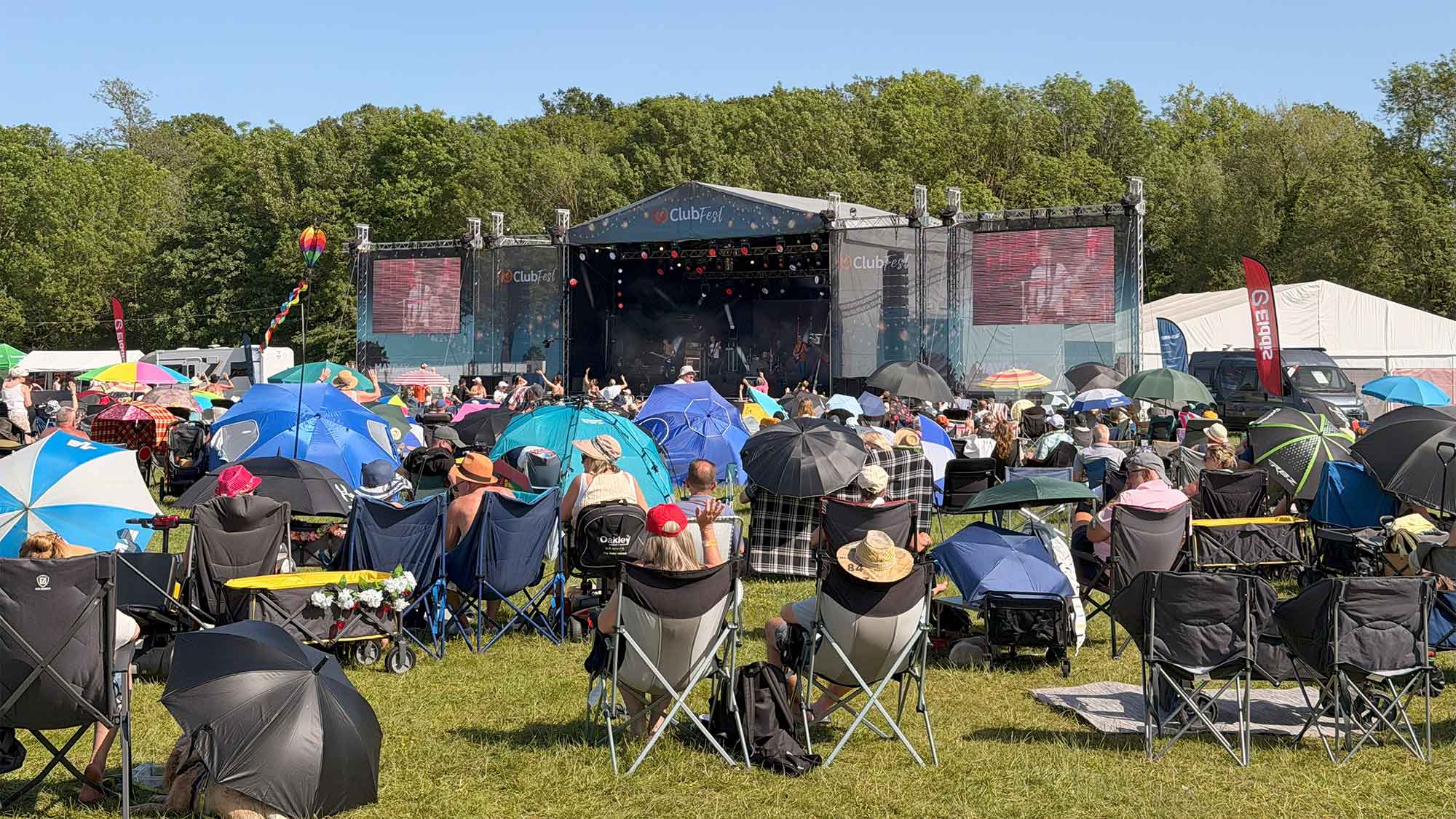 A large crowd sitting on camping chairs on the green grass with a number of colourful umbrellas shading people from the hot sunshine. Facing a large stage at ClubFest 2026. The tall green trees rising up in the distance under a bright blue sky.