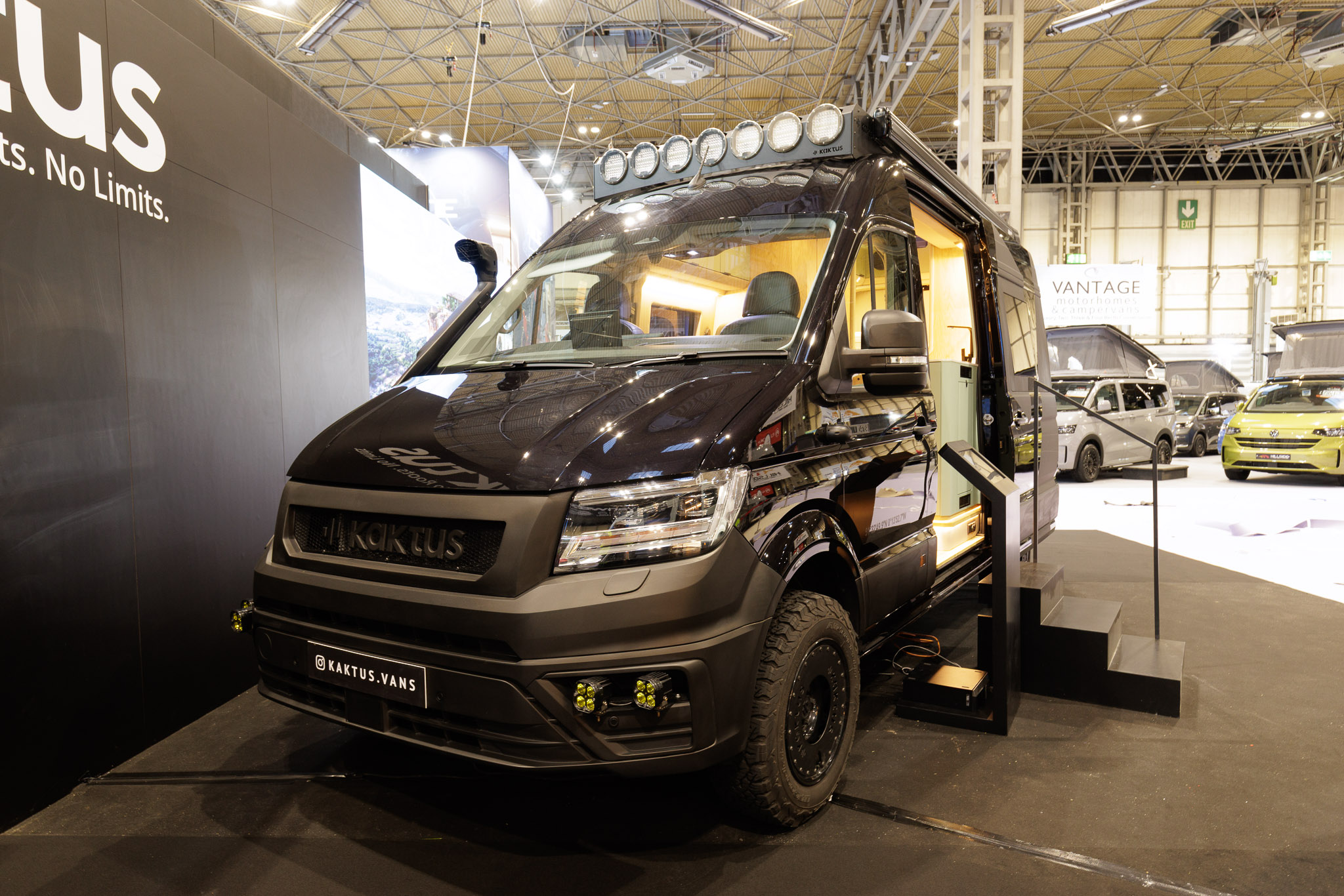 The Kaktus Adventure Vans Dune is black, its sliding door is open and there are three steps to gain easy access.  On the cab roof there are seven spotlights.