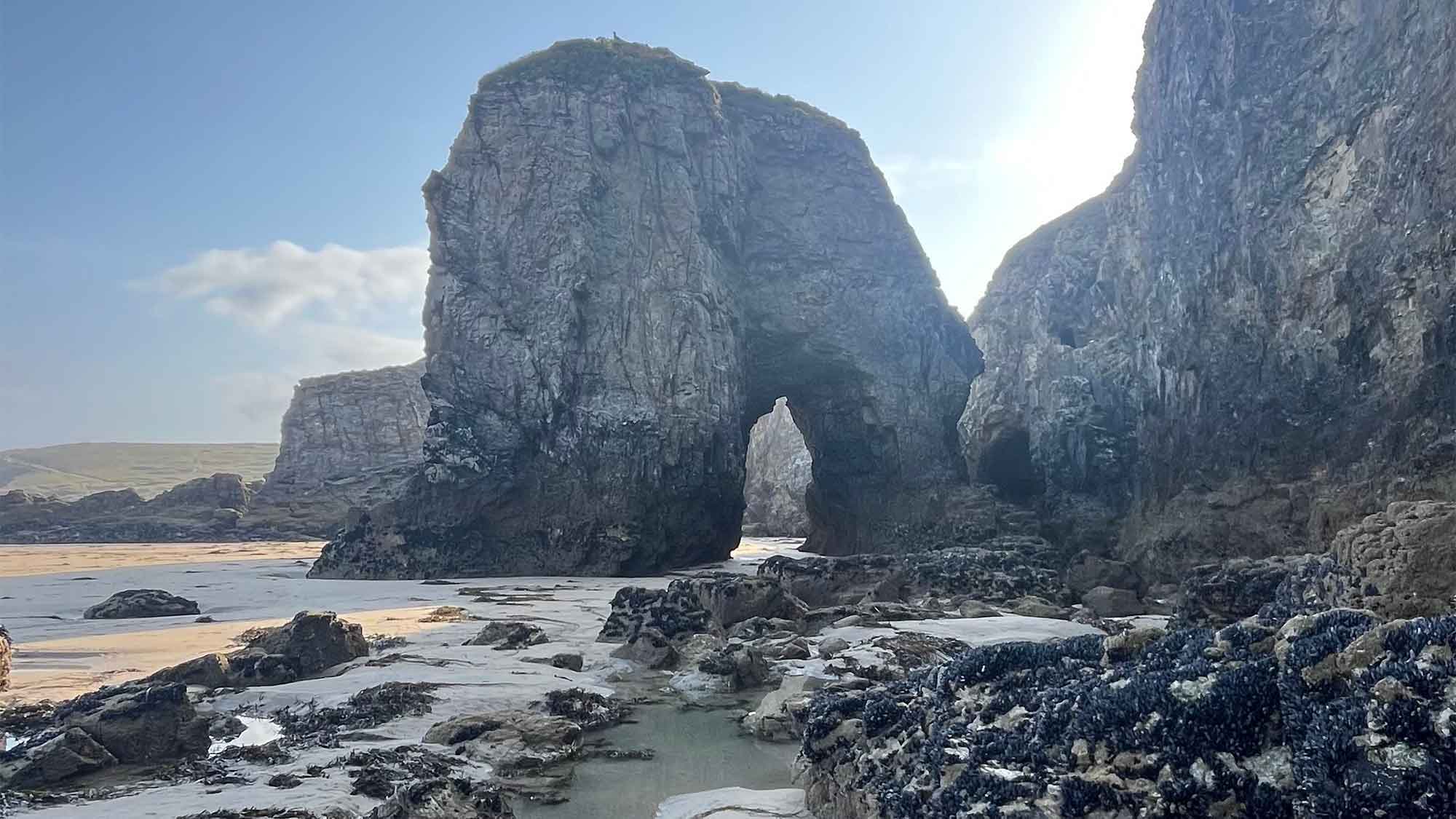 Arch of rock beside the cliffs with sand a rock pools in front under a blue sky