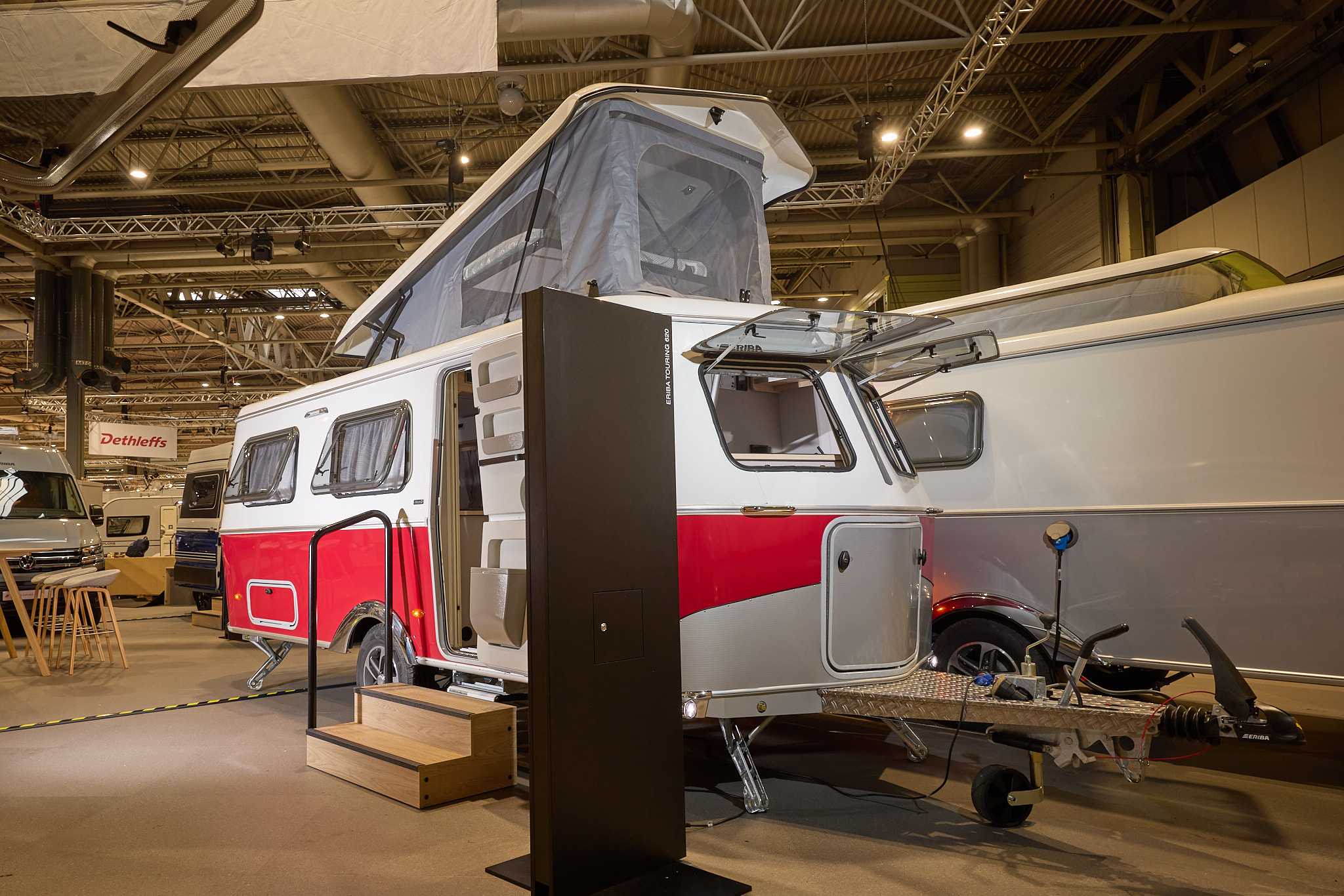 The classically styled Eriba Touring 620 Tango Red Edition has a two tone layout in red and white.  Its campervan style pop top roof is open as is its entrance door.  There are two wooden steps to gain easy access.