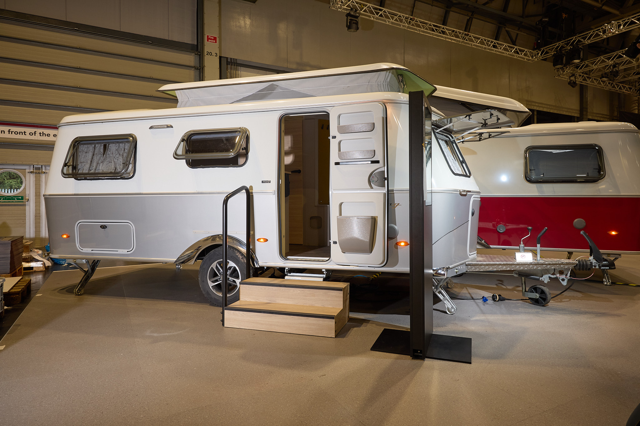 The classically styled Eriba Touring 630 Urban has a two tone layout in silver and white.  Its pop top roof is open as is its entrance door.  There are two wooden steps to gain easy access.