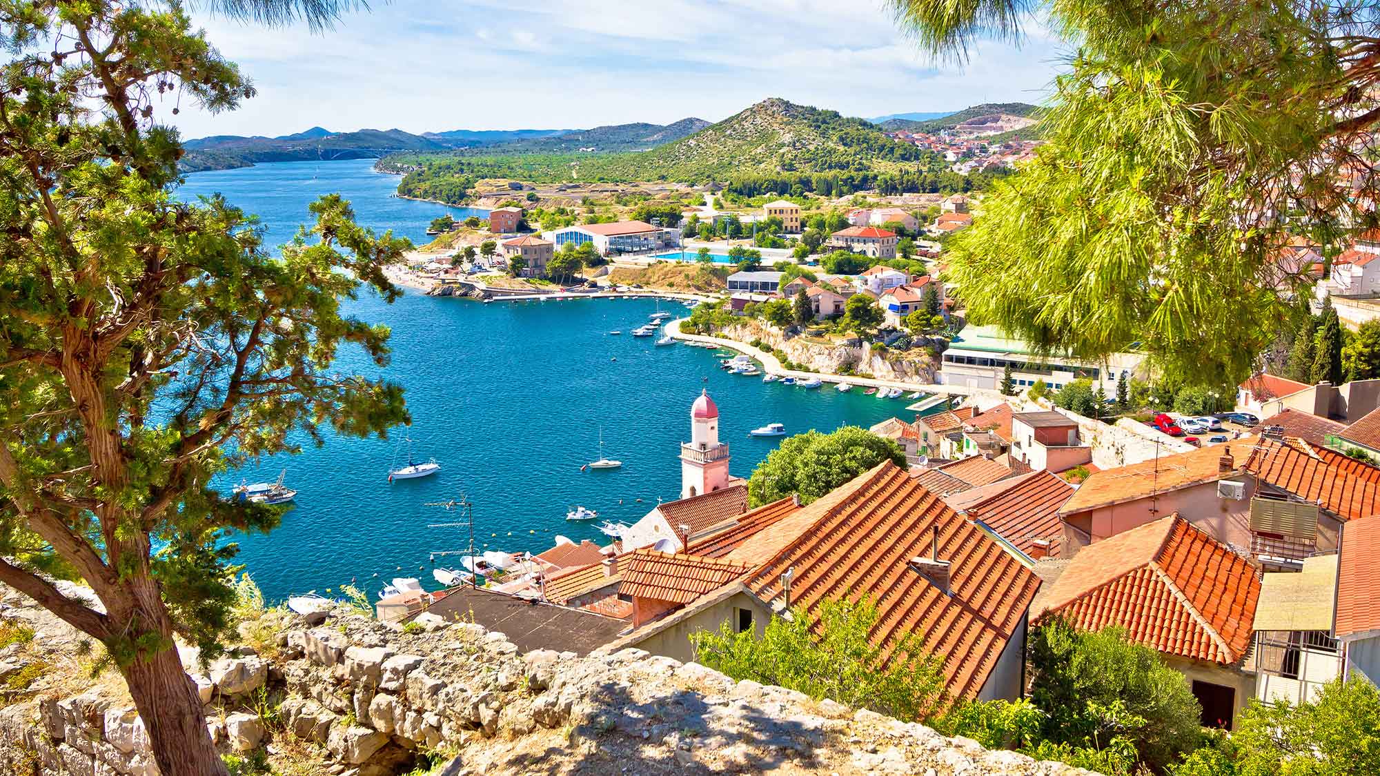 A view of Sibenik, Croatia. Looking down from an elevated viewpoint framed by trees looking over the rooftops leading down to the stunning sea
