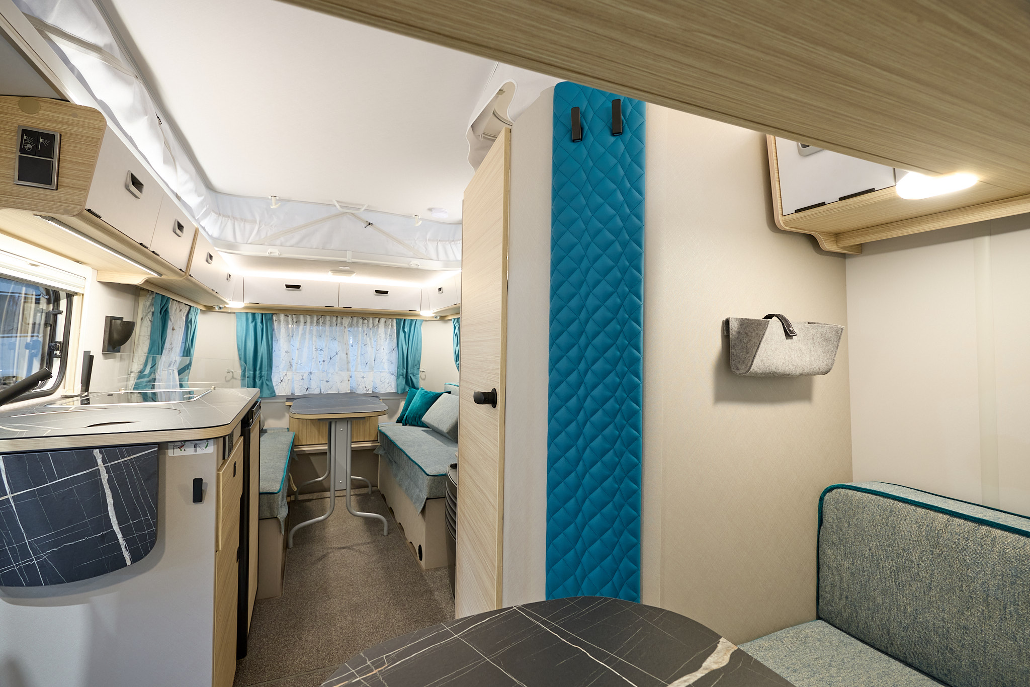 Eriba Touring 540 Harbour Blue Edition’s interior has grey upholstery and its furniture is pale wood.  There are sofas with tables at the front and the rear.  There is a blue panel with two black hooks to the right.