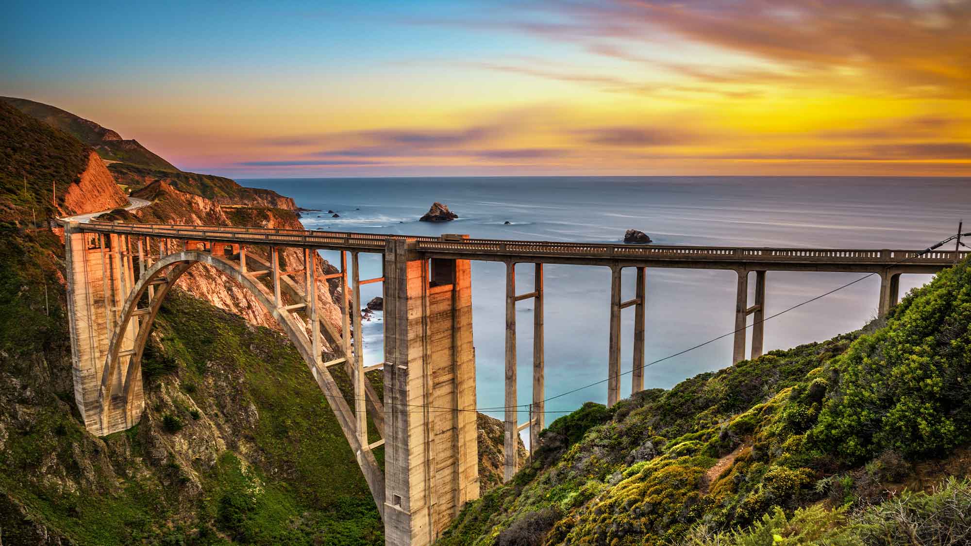 Very tall bridge structure over a valley with the ocean in the background under a beautiful sunset