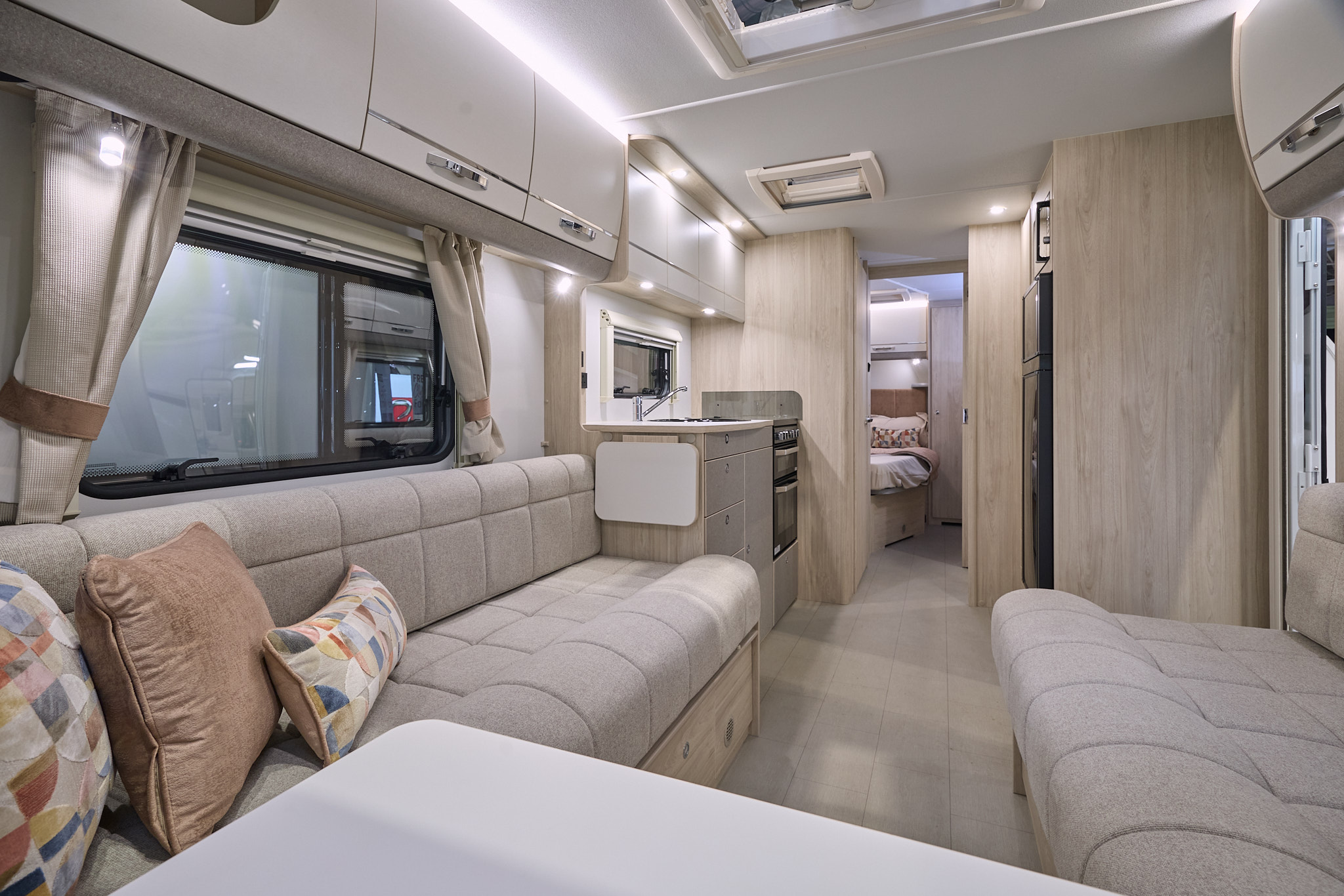 Elddis Avanté 868’s interior has beige upholstery with pale wooden furniture.  The kitchen is in the middle and the bedroom door is open revealing a French transverse fixed bed.