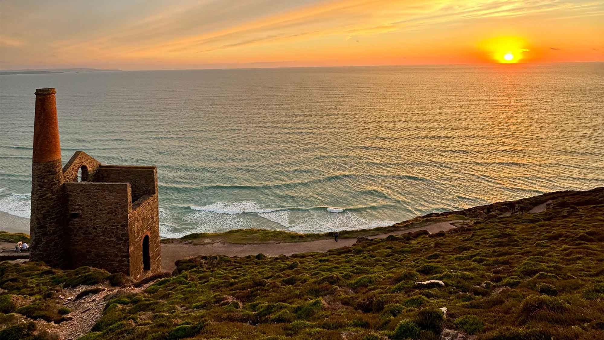 Sunset over the sea looking out from the grassy hill with an old stone building of Wheal Coates next to the shoreline