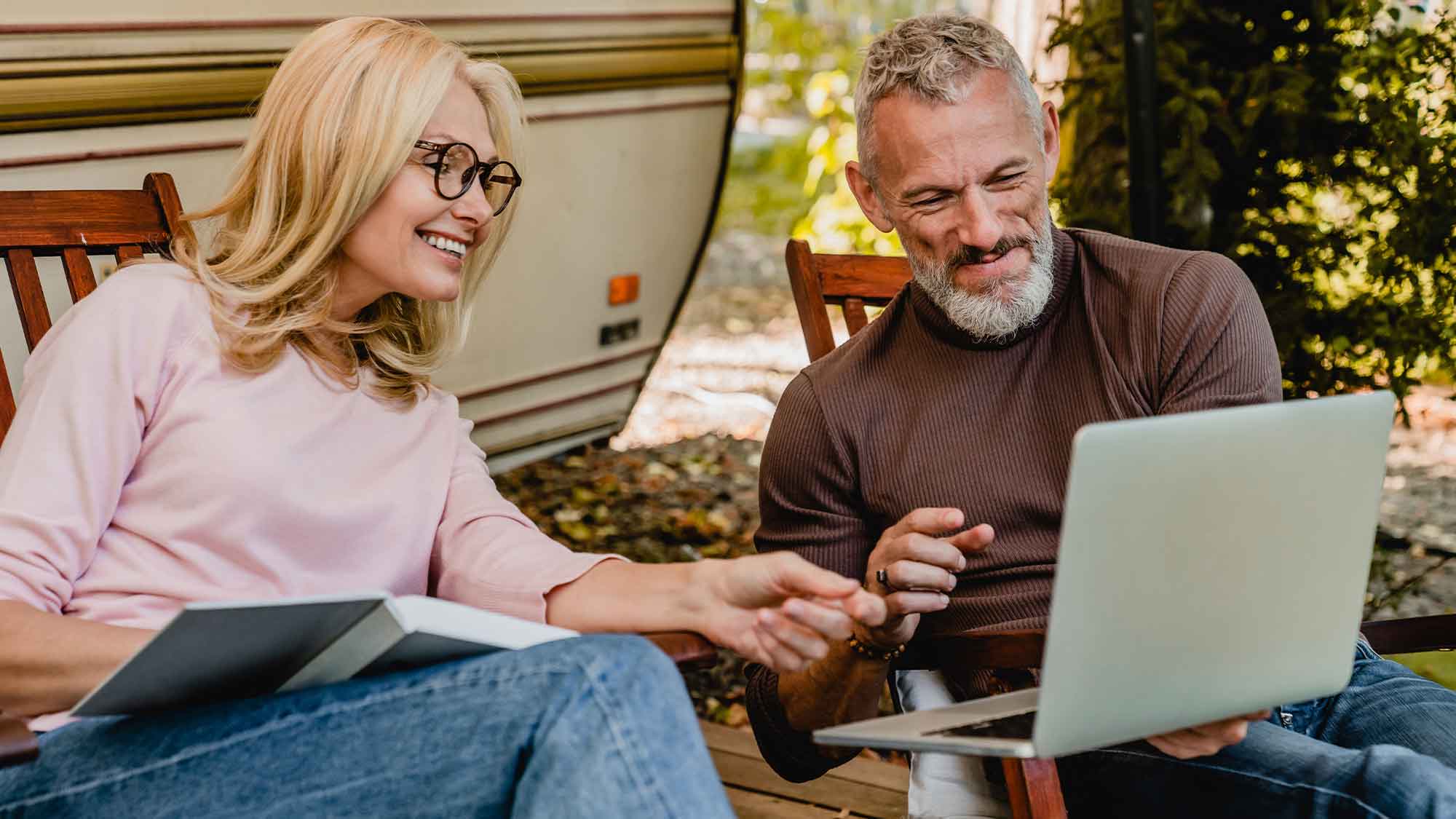 A man and a women both smiling looking at laptop sitting on chair with a caravan in the background