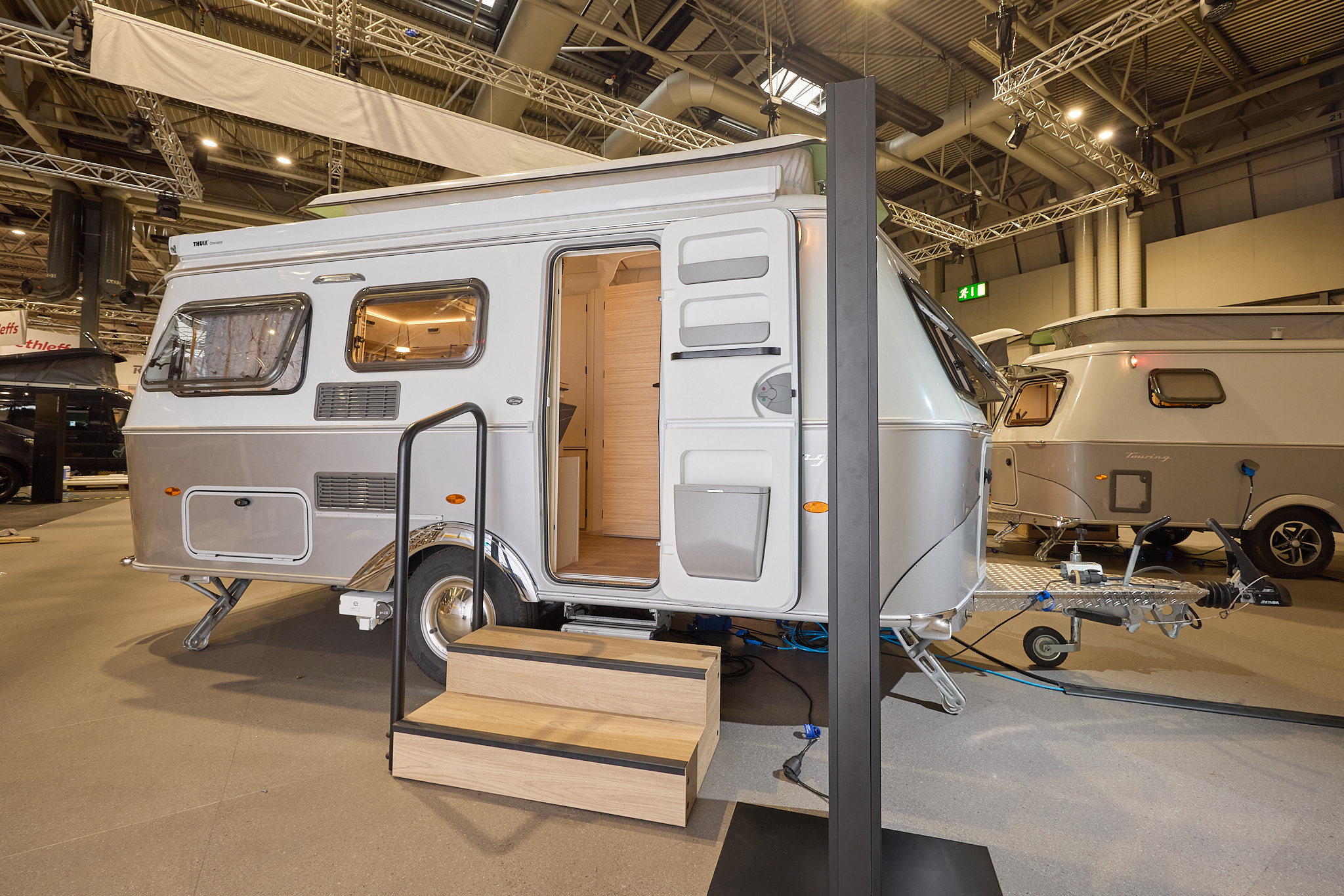 The classically styled Eriba Touring 530 Urban has a two tone layout in silver and white.  Its pop top roof is open as is its entrance door.  There are two wooden steps to gain easy access.