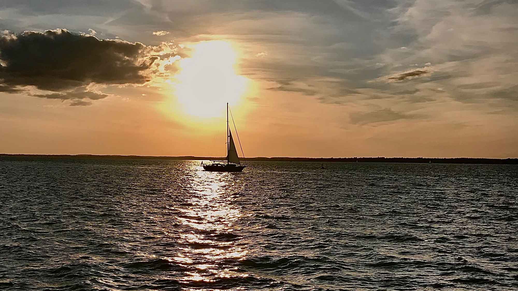 a small sailing boat out in the bay with the setting sun at the top of the mast