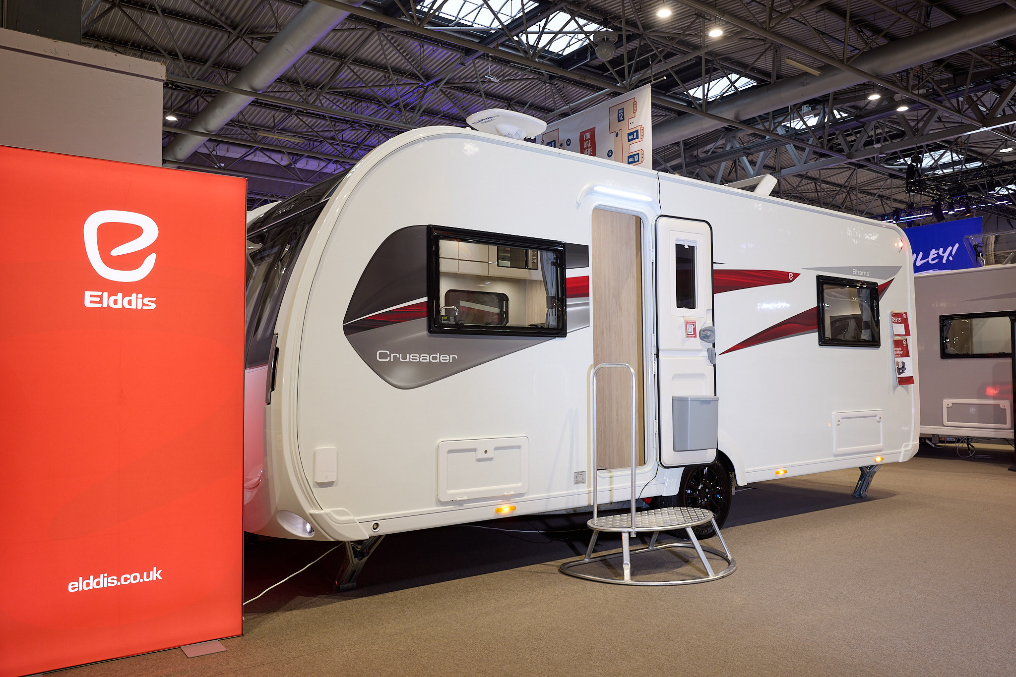The Elddis Crusader Shamal has a white exterior with grey, red and black decals.  The entrance door is open and there is a metal step to gain easy access.  There is a large Elddis red banner with their website address next to it.