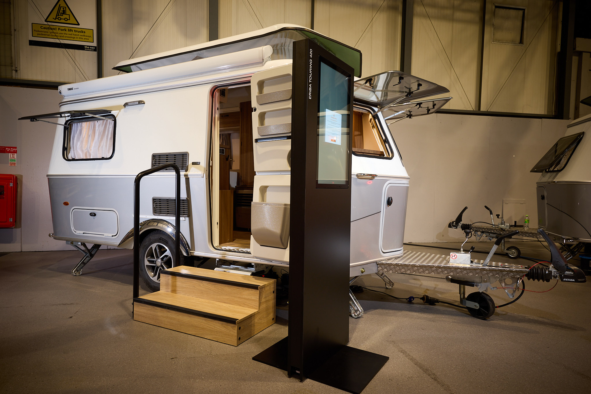 The classically styled Eriba Touring 420 Legend has a two tone layout in silver and white.  Its pop top roof is open as is its entrance door.  There are two wooden steps to gain easy access.